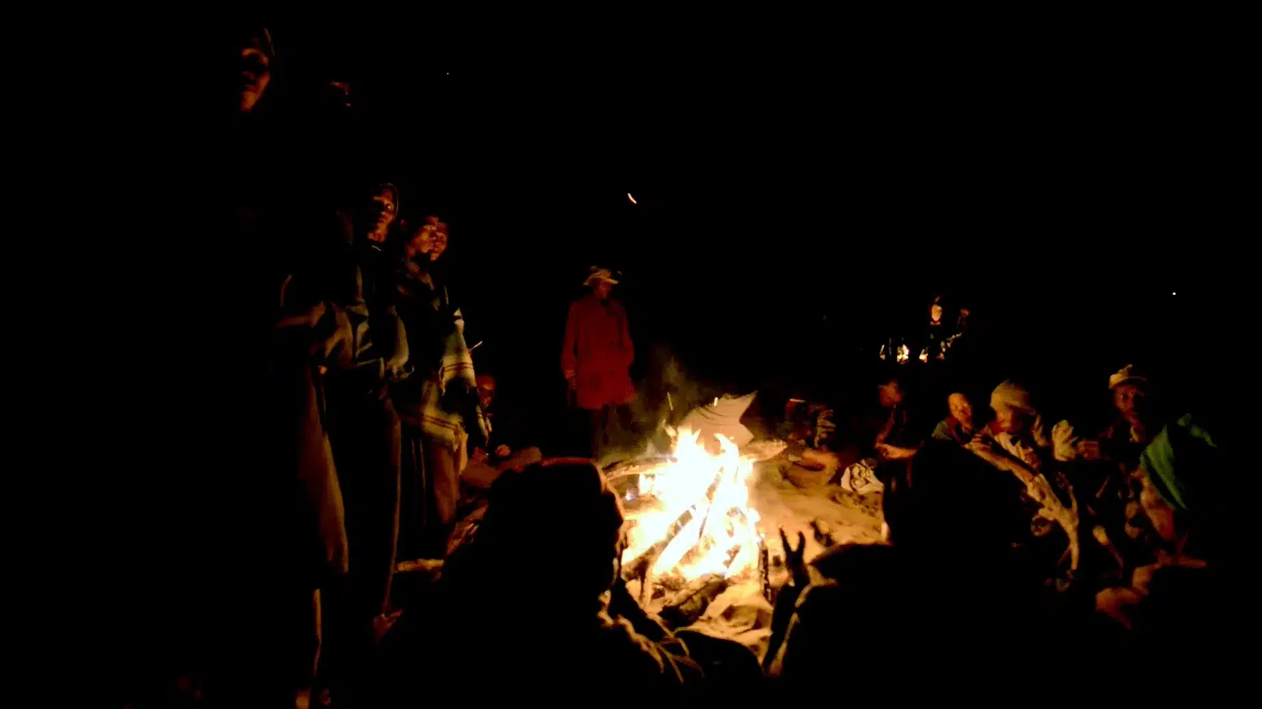 A group of people sit around a campfire at night, with soft glowing light illuminating their faces. Some are standing and some are sitting, wrapped in warm clothing, while the flickering flames create a cozy, communal atmosphere against the dark background.