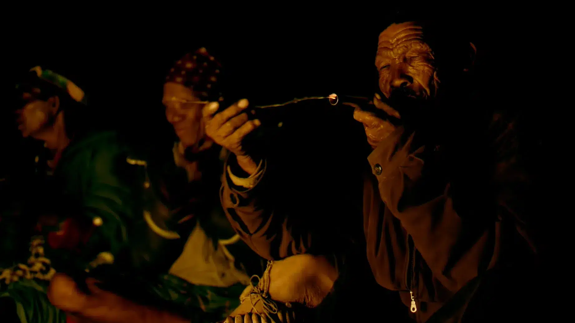 A group of people sit around a campfire at night. An older person in the foreground is holding and smoking a pipe, while others in the background are engaged in conversation, illuminated by the fire's warm glow. The overall atmosphere feels relaxed and communal.