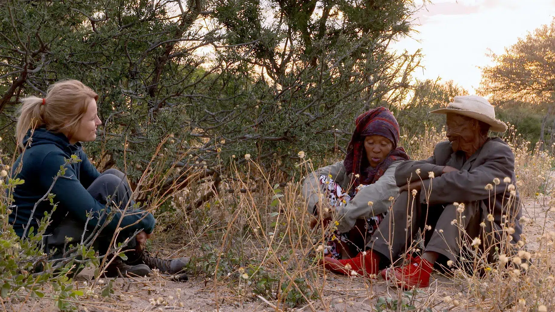 Three individuals are seated and conversing in a natural setting surrounded by dry grasses and bushes. One person appears to be showing items, possibly gemstones, to the others. The sun is setting in the background, casting a warm glow over the scene.
