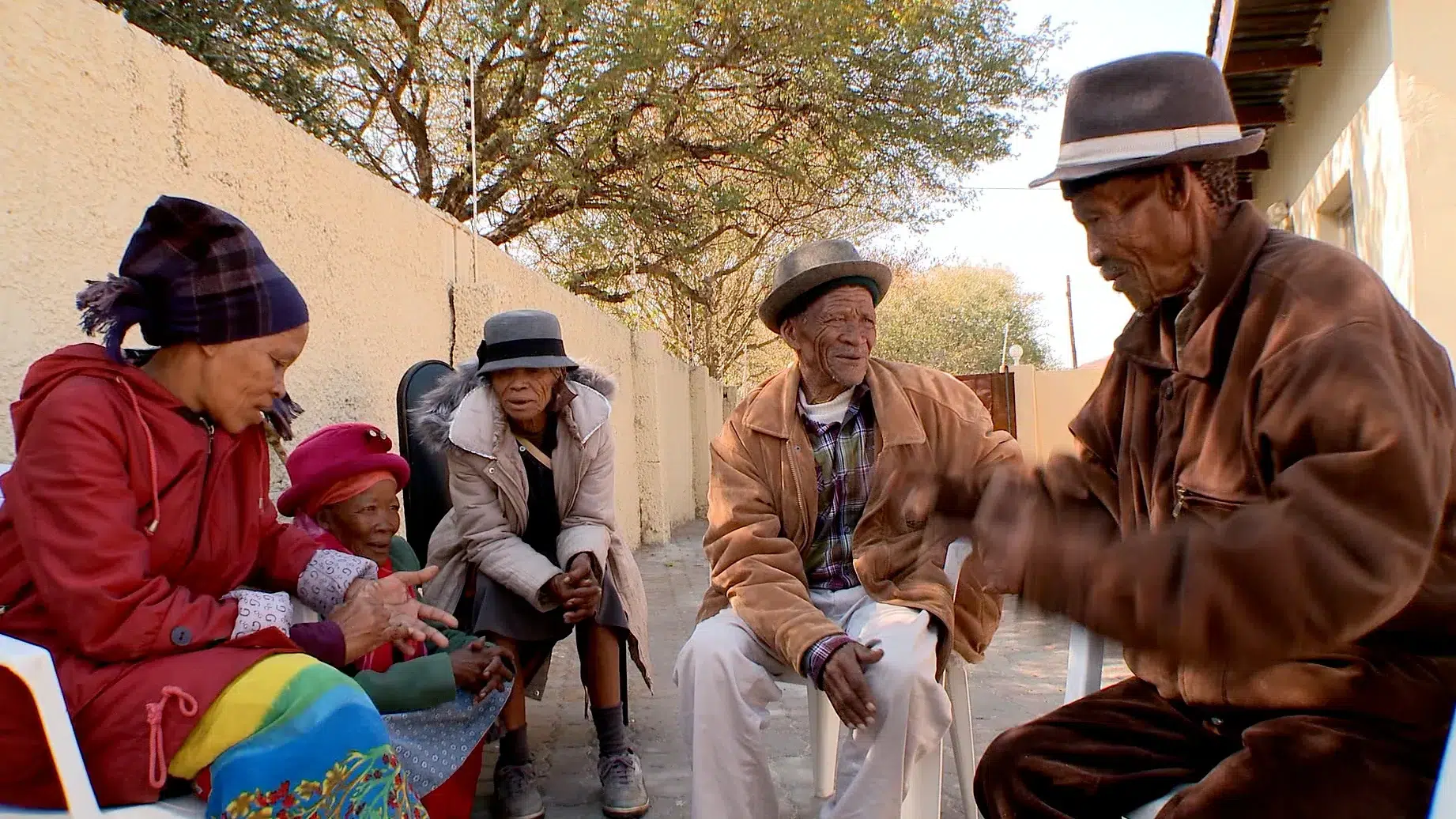 A group of five elderly people sit outside on a sunny day, engaging in conversation. They are dressed warmly in coats and hats. The background shows a wall, trees, and a partial view of a building.
