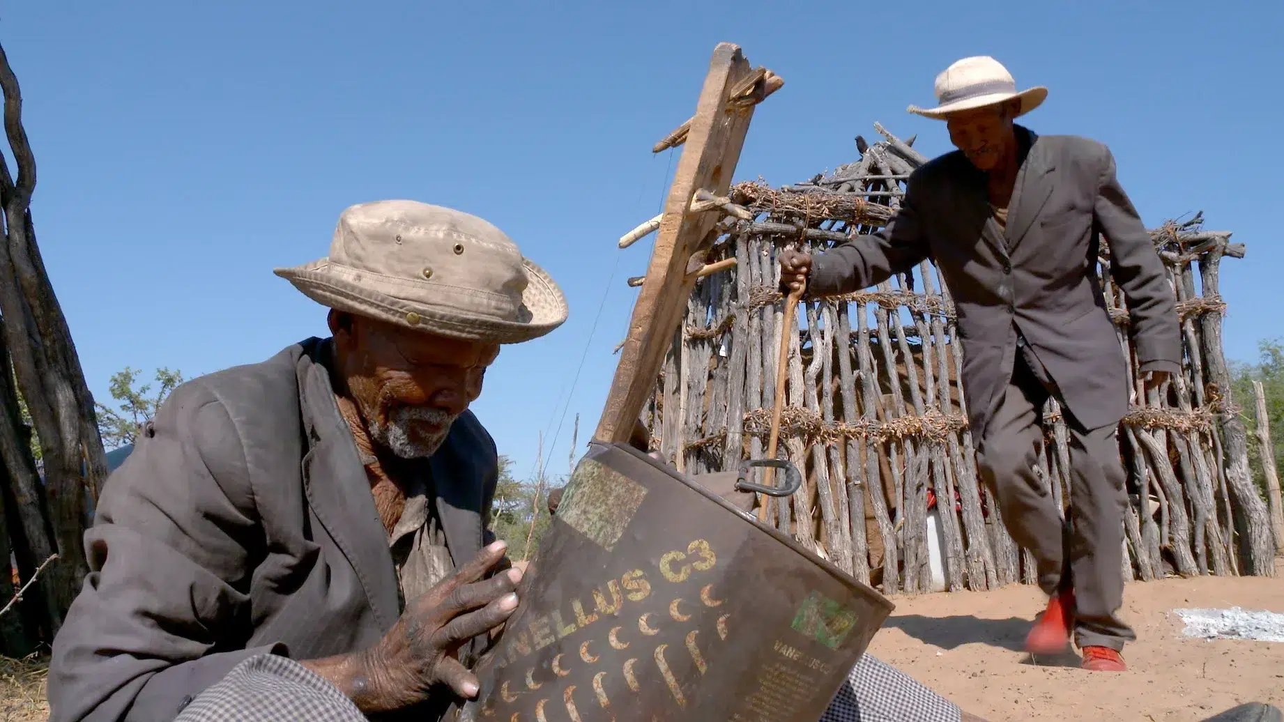 Two elderly men in worn suits are outdoors; one is kneeling and examining a metal canister labeled "HELLUS C3," while the other, walking with a wooden cane, approaches a rustic, wooden structure in the background under a clear blue sky.