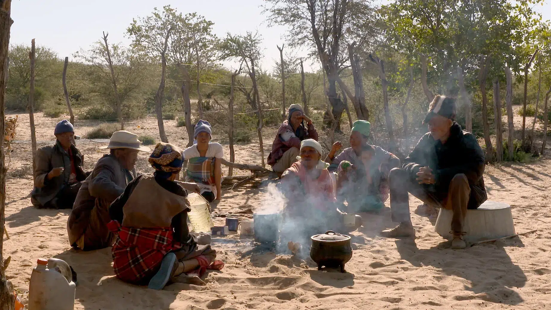A group of people are seated on the sandy ground around a small campfire in an outdoor setting. Various cooking pots and utensils are placed near the fire. Trees and a rustic wooden fence are visible in the background. Everyone appears to be engaged in conversation.