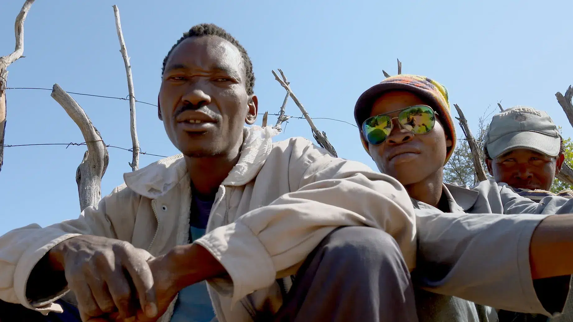 Three men sit outdoors in a rural area. The man in the foreground wears a light jacket, the man in the middle wears reflective sunglasses and a colorful hat, and the man in the background wears a cap, blending into the surroundings of wooden sticks and clear blue sky.