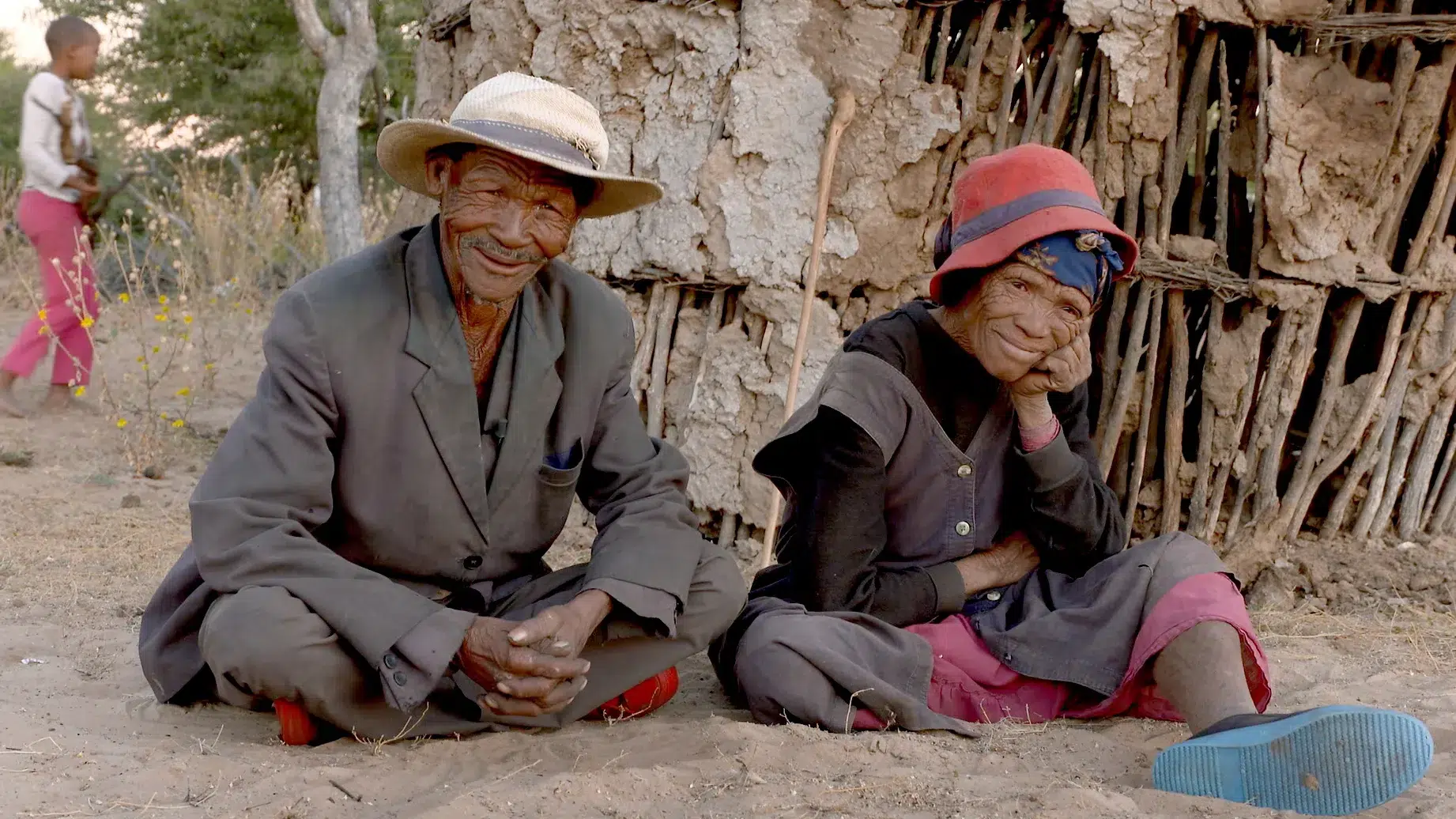 Two elderly individuals smile while sitting in the dirt outside. The man wears a suit jacket, dark pants, and a white hat. The woman wears a dark dress, pink pants, and a red hat, resting her head on her hand. An adobe structure and another person are visible in the background.