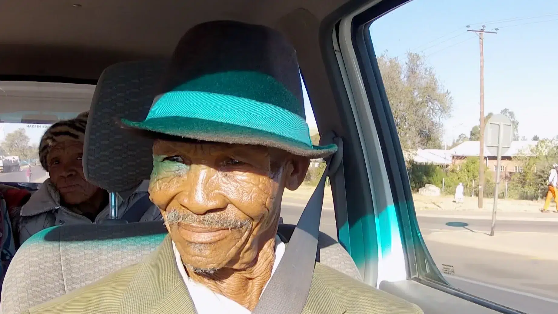 An elderly man wearing a green and black hat and brown blazer sits in the passenger seat of a car, smiling. Another person in a striped hat is seated behind him. The car is parked on a sunlit street with trees and utility poles visible outside the window.