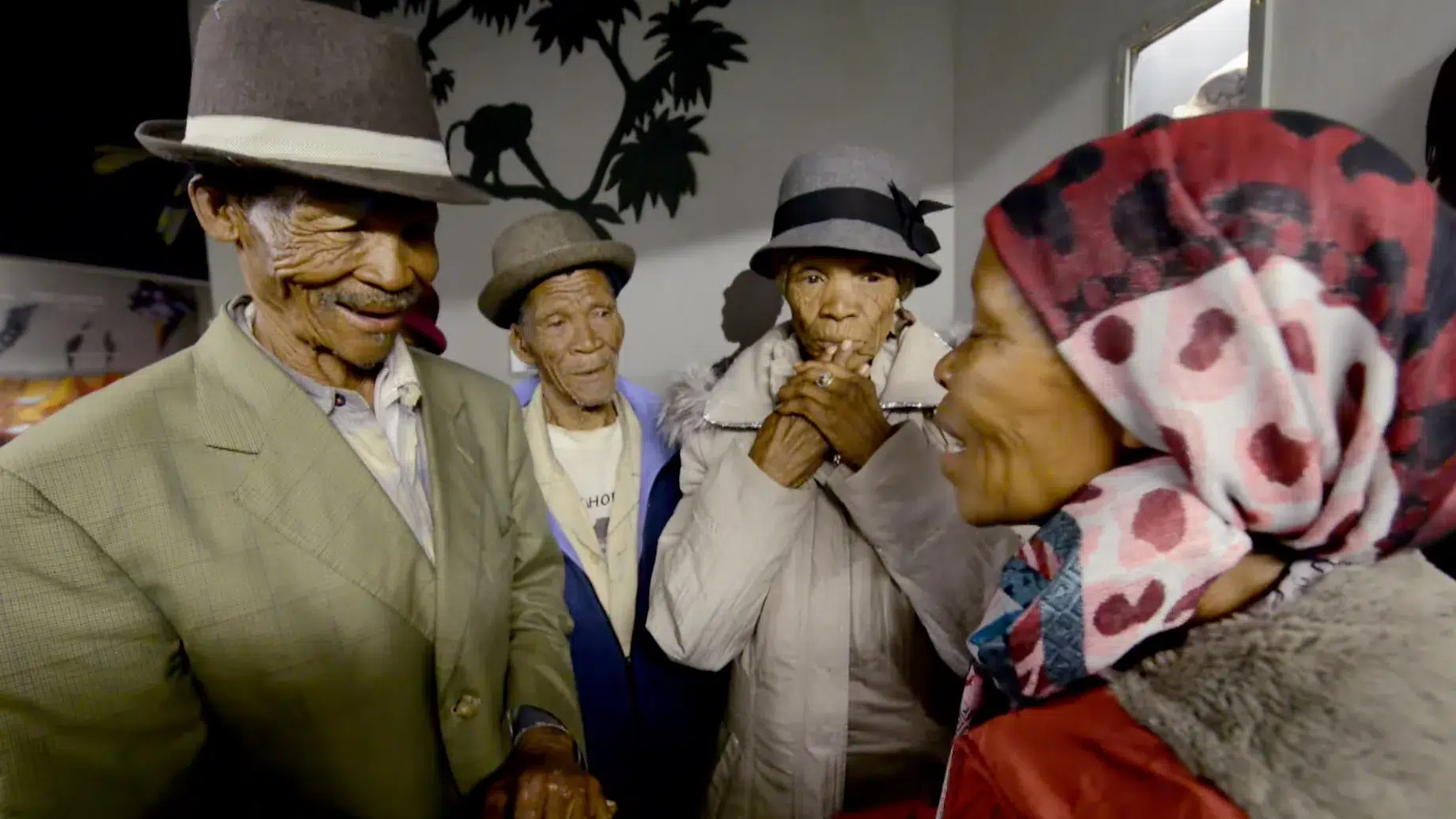 A group of older people dressed in hats and coats, engaged in conversation and sharing smiles indoors. One person wears a spotted headscarf, while others sport wide-brimmed hats. The background features a wall mural with silhouettes of plants and animals.