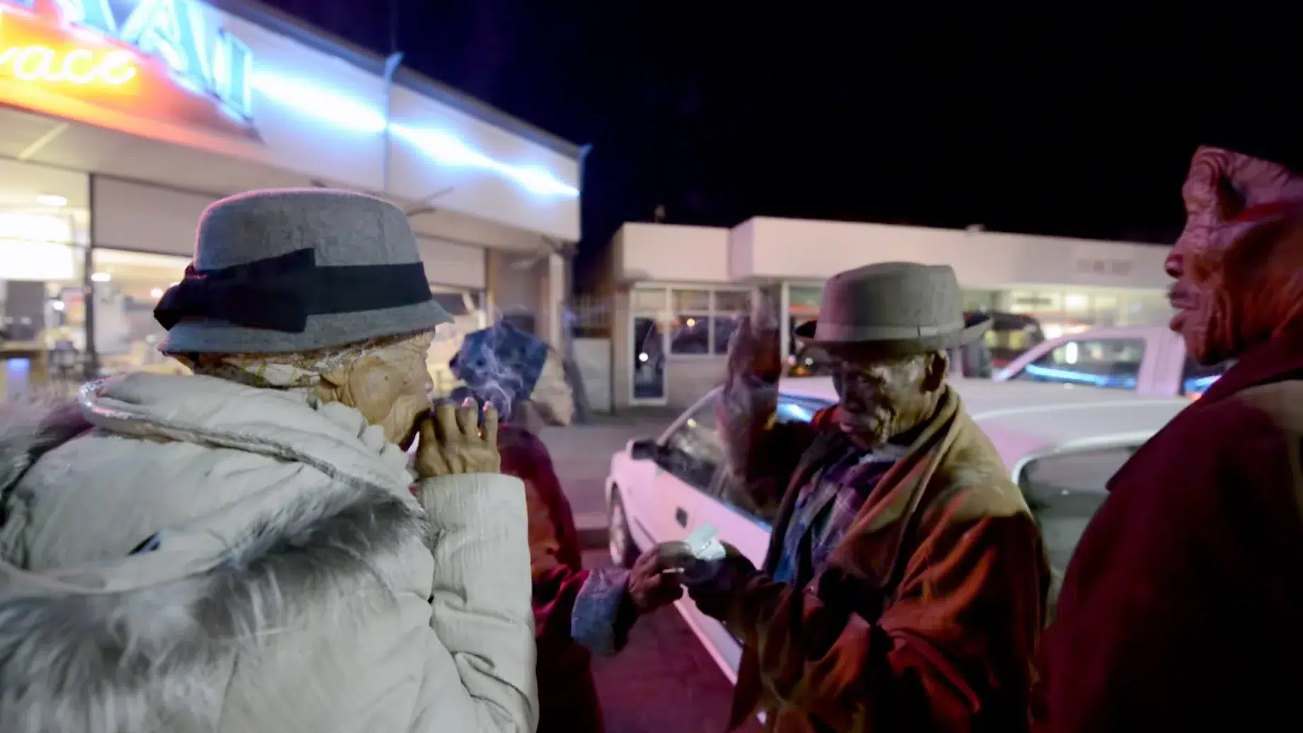 Four older adults, wearing winter coats and hats, gather and converse outside at night. They stand in the foreground in front of a brightly lit building with a neon sign, and a white car is parked nearby. One person appears to be holding something in their hands.