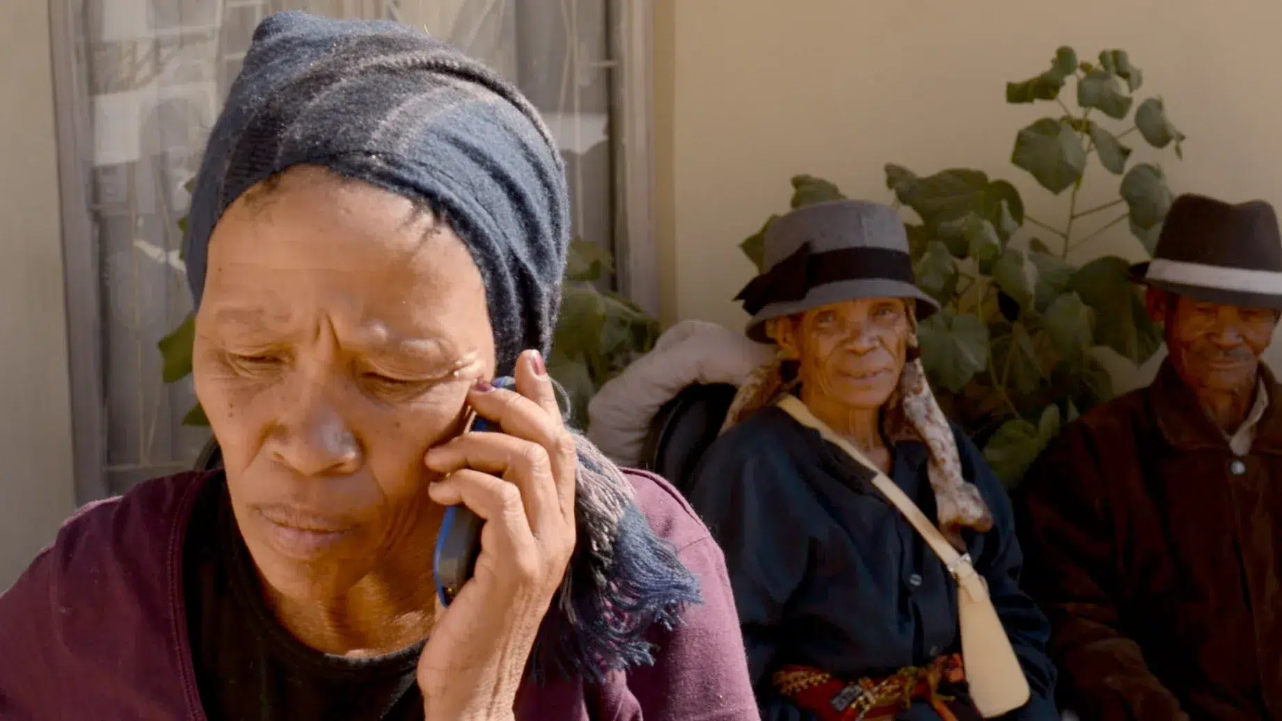 A close-up of an elderly woman talking on a mobile phone, wearing a headscarf. In the background, two other elderly individuals, also wearing headscarves, are seated, smiling, and observing the scene. There is a leafy plant behind them.