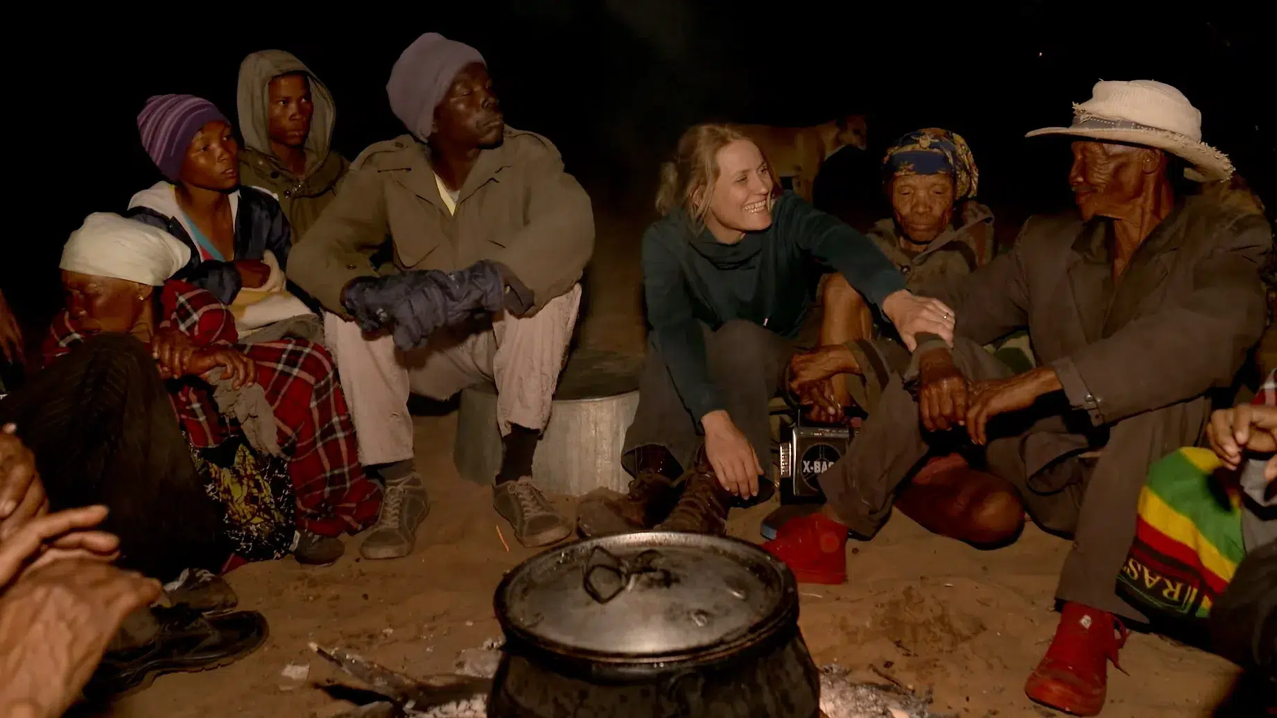 A group of people sit in a circle around a fire, with a large pot placed on the ground in the center. They are dressed warmly, and appear to be engaging in conversation and sharing a communal moment in an outdoor setting at night.