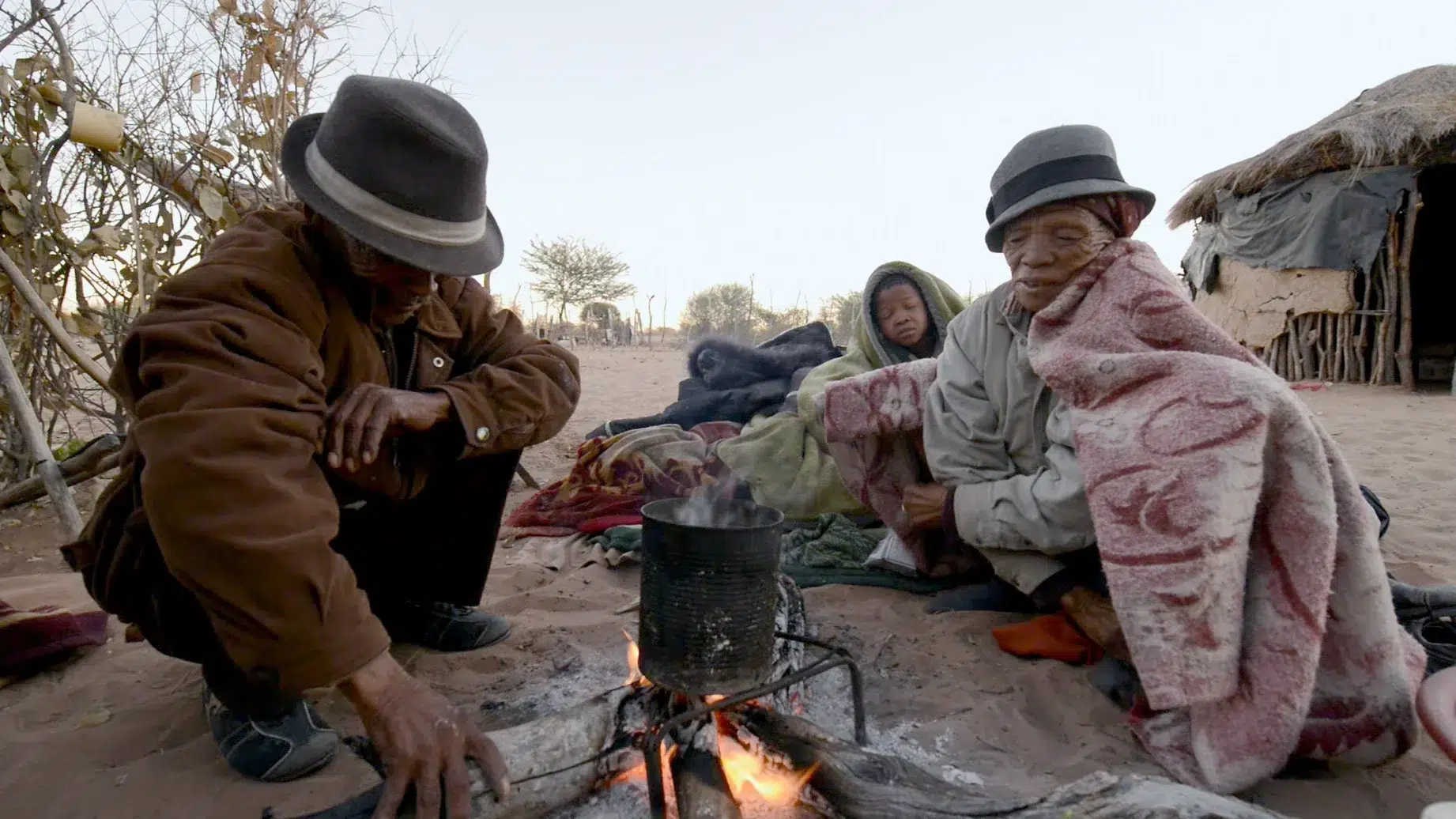 Three individuals sit around a small fire in a sandy outdoor setting. They are bundled up in warm clothing and blankets. A pot is placed on the fire. Straw huts and sparse vegetation are visible in the background. The scene appears to be in a rural area.