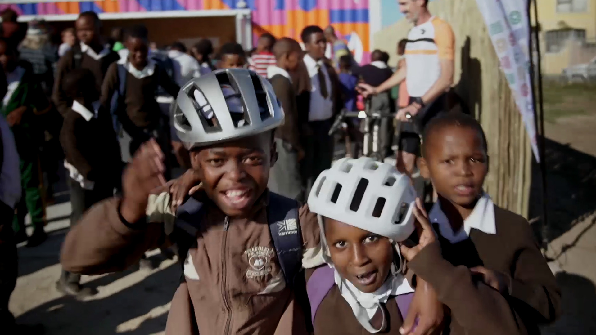 Three children wearing helmets smile and pose for the camera while a crowd of other children and a few adults gather in the background. The scene appears lively and energetic.