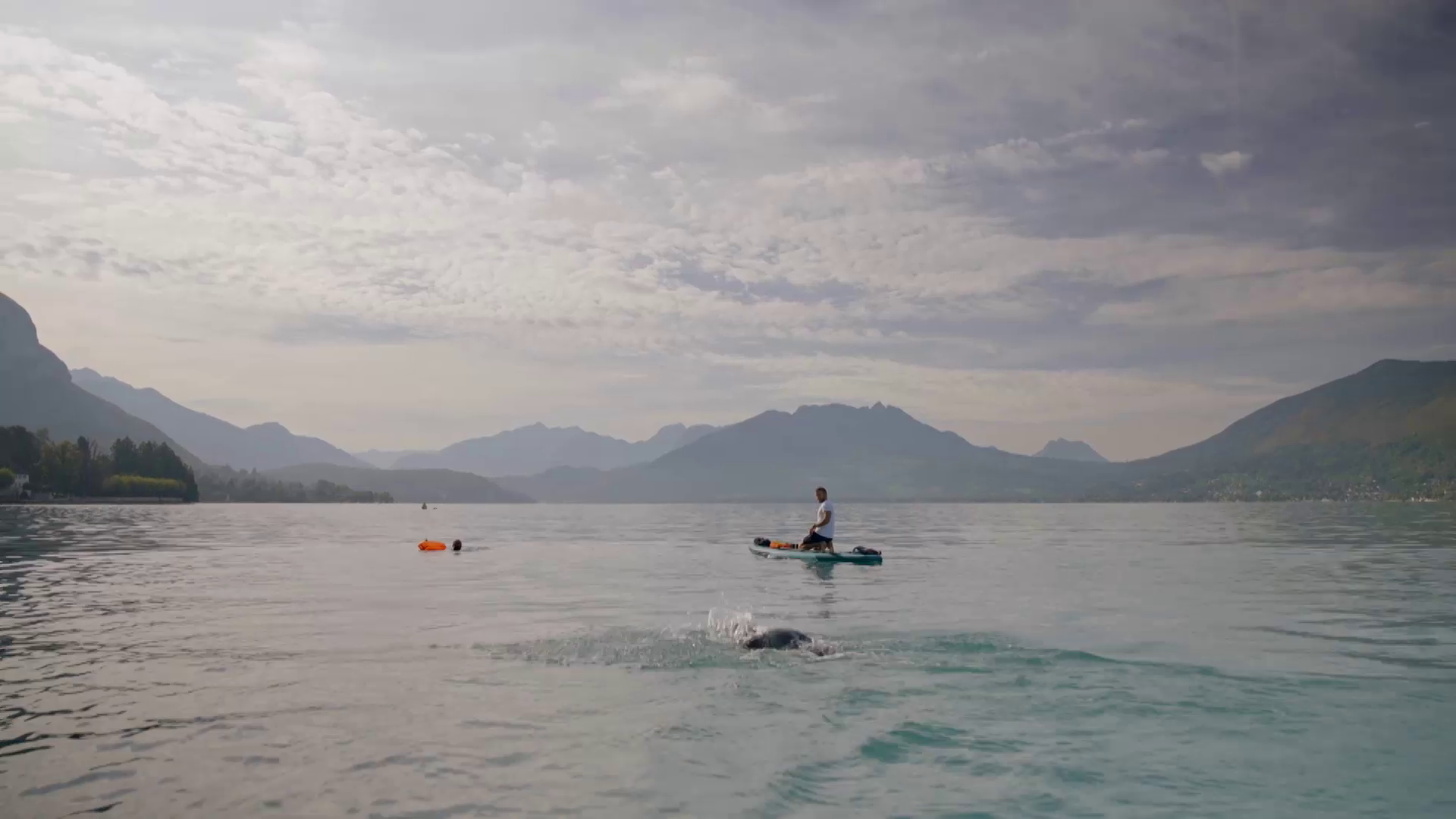 A serene lake surrounded by mountains under a partly cloudy sky. A person on a paddleboard is paddling in the distance while another person swims in the foreground. An orange buoy floats on the water's surface.