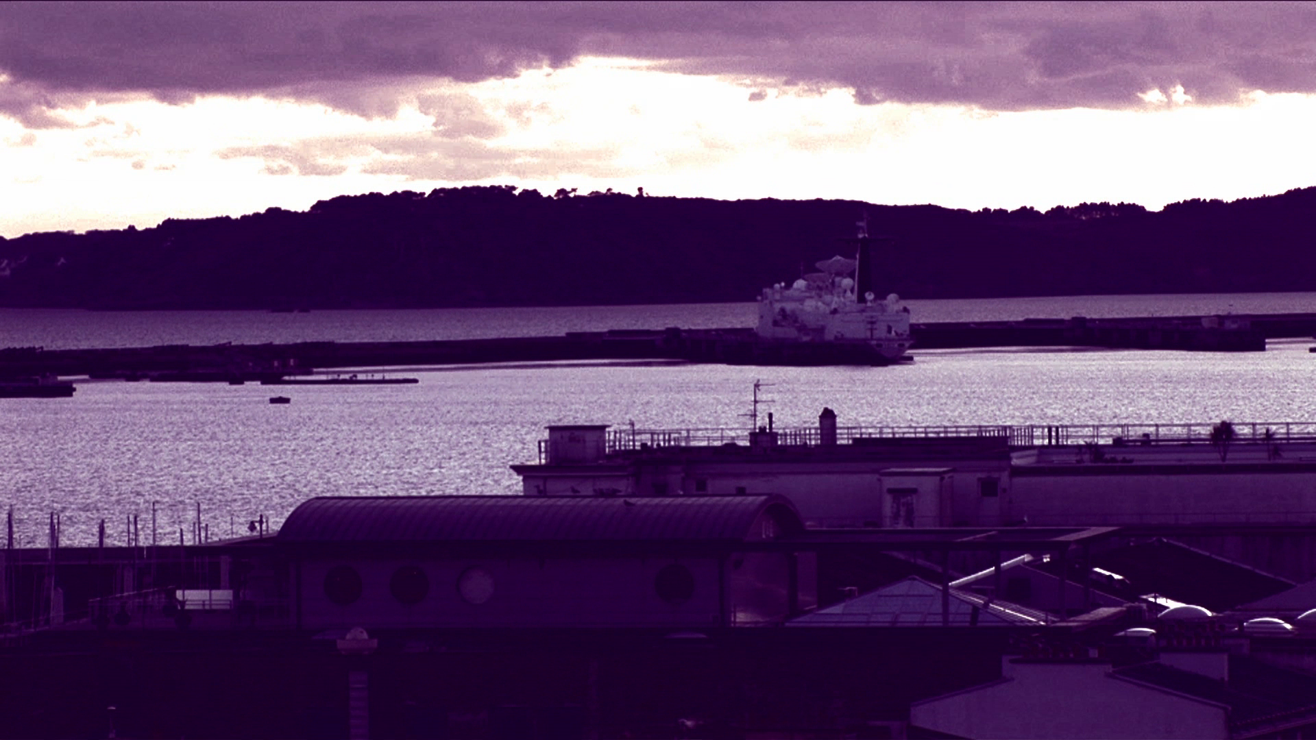 •10 Xpress2Brest_Film_Kate_Thompson_Gorry A serene waterfront scene at dusk, with a large ship docked near a shoreline. The background features silhouetted hills under a cloudy sky with shades of purple and pink, reflecting off the calm water. Several buildings are visible in the foreground.