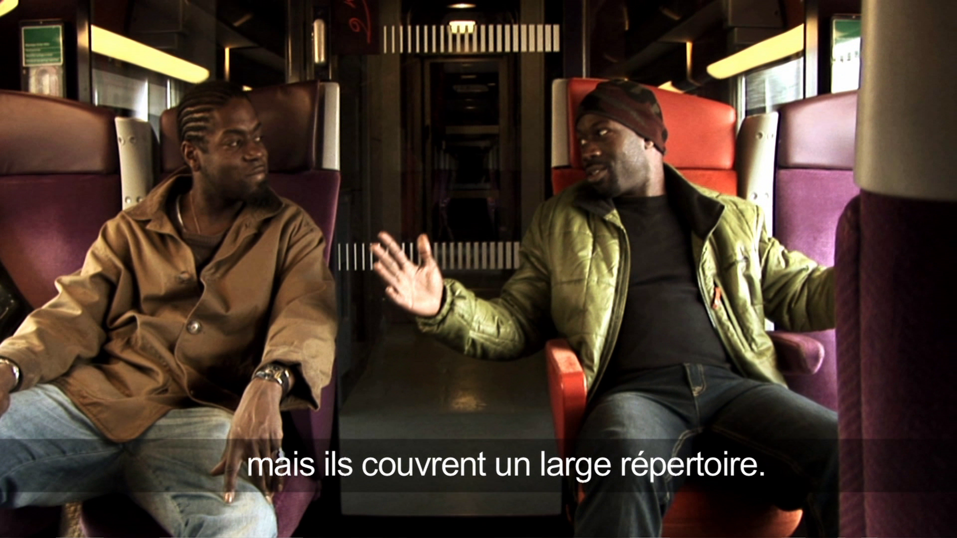 •11 Xpress2Brest_Film_Kate_Thompson_Gorry Two men sit facing each other on a train. One is wearing a brown jacket and jeans, while the other has on a green jacket and a beanie. They appear to be in conversation. French text on the screen reads, "mais ils couvrent un large répertoire," translating to "but they cover a wide repertoire.