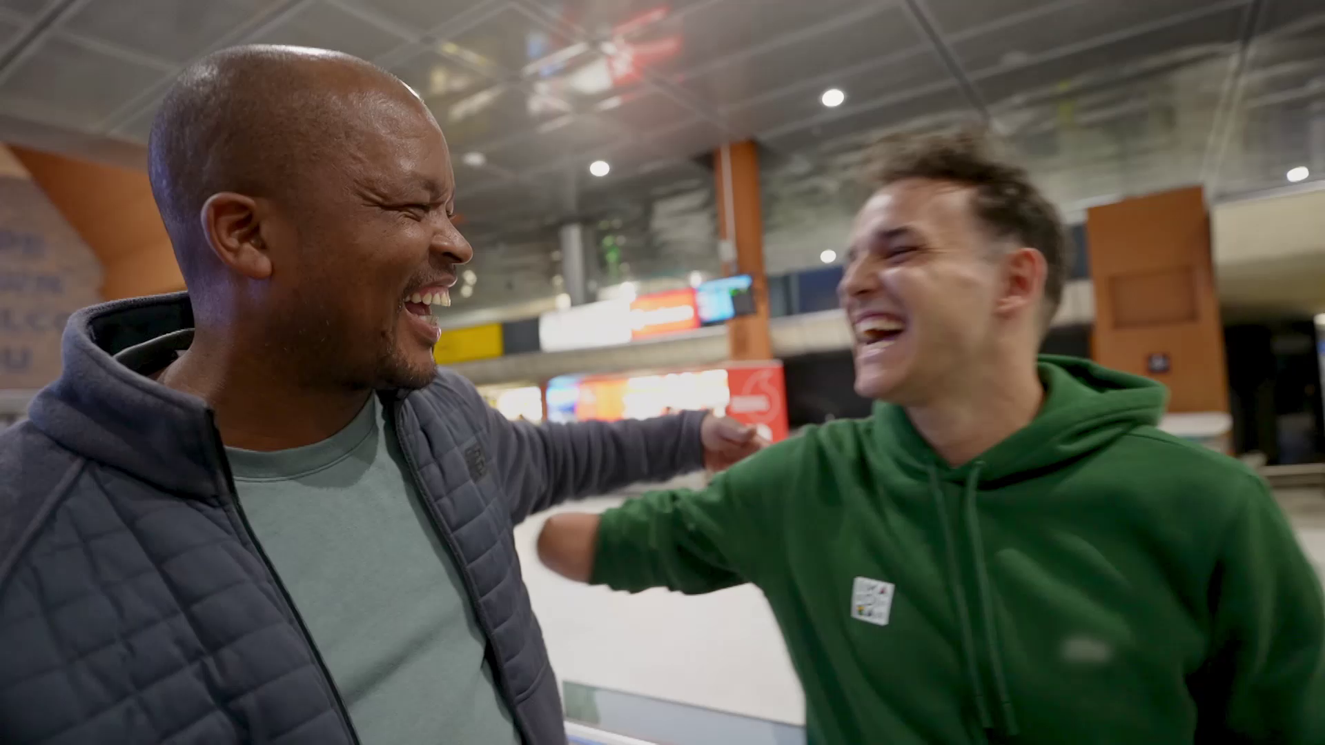 Two men are standing inside a brightly lit building, smiling and laughing while embracing each other. One man is wearing a dark jacket and a green shirt, and the other man is in a green hoodie. They appear to be sharing a joyful moment.