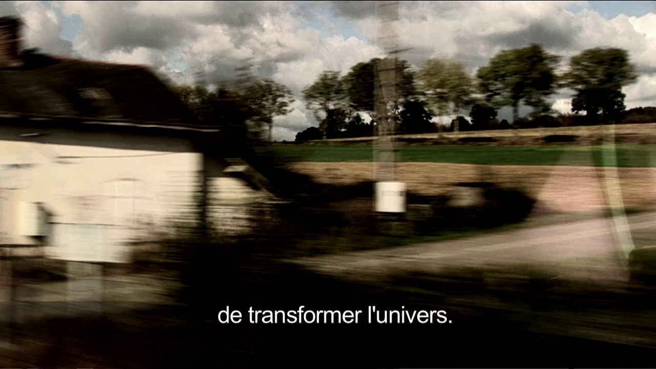 •12 Xpress2Brest_Film_Kate_Thompson_Gorry A blurred scene of a countryside with a house on the left, fields, and trees in the background under a partly cloudy sky. French text at the bottom reads, "de transformer l'univers" which translates to "to transform the universe.