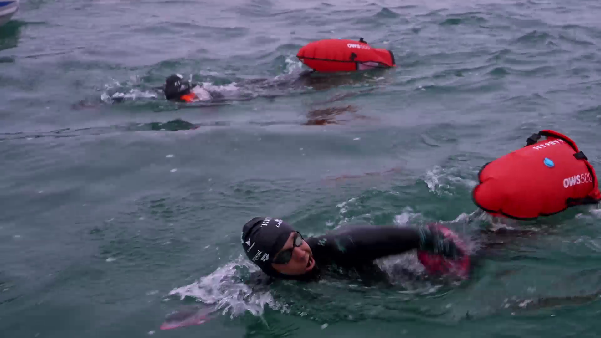 Two swimmers in wetsuits are swimming in open water with red buoy floats attached to them. They are surrounded by choppy waves, and one swimmer is in the foreground while the other is slightly behind. The scene appears to be part of The Madiba Challenge, an intense open-water race or training session.