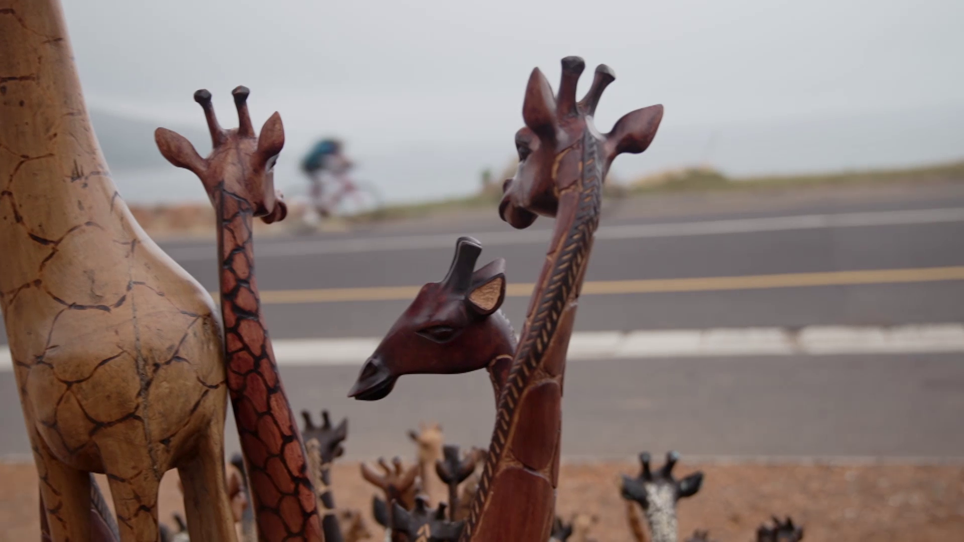 Wooden giraffe statues of various sizes are displayed outdoors near a road. In the blurred background, a few cyclists can be seen riding along the road with a cloudy sky overhead. The giraffes have intricate carvings and different shades of brown.