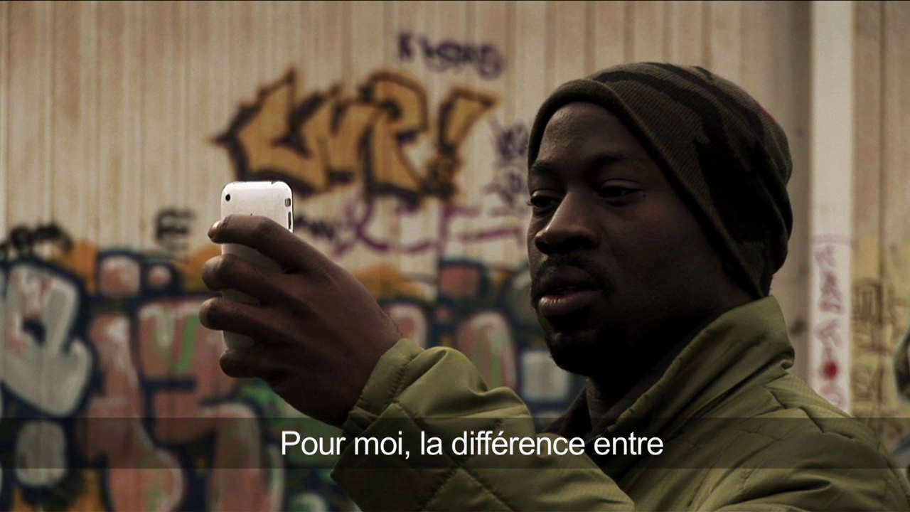 •14 Xpress2Brest_Film_Kate_Thompson_Gorry A person wearing a green jacket and dark-colored beanie is holding up a smartphone, taking a photo or video. There is graffiti art on the wall behind them. The text at the bottom of the image in French reads, "Pour moi, la différence entre.