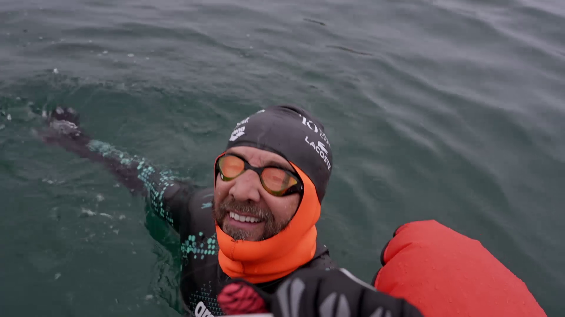 A person wearing a wetsuit, swim cap, and orange neck covering is swimming in open water. They have goggles on and appear to be reaching out with their arm, possibly toward the camera. The water around them is calm.