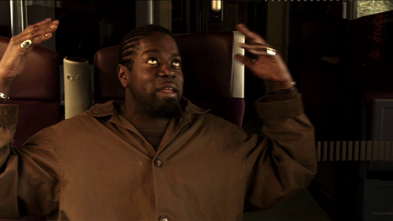 •15 Xpress2Brest_Film_Kate_Thompson_Gorry A person with braided hair, wearing a brown button-up jacket and a ring on a finger, is sitting in a chair and gesturing with both hands raised, as if emphasizing a point or expressing frustration. The background appears to be an indoor setting with dim lighting.