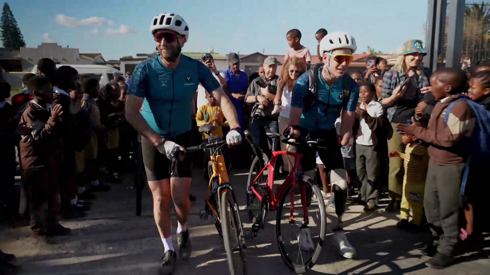 Two cyclists wearing matching blue jerseys and white helmets walk their bicycles through a crowd of cheering children and adults. The scene appears joyful and celebratory, with people clapping and taking photos.