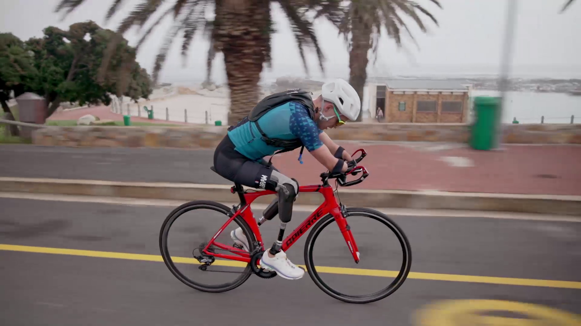 A cyclist in a blue and green jersey and black cycling shorts rides a red road bike on a paved road. They wear a white helmet and carry a small backpack. Palm trees and a sandy beach can be seen in the background on an overcast day.