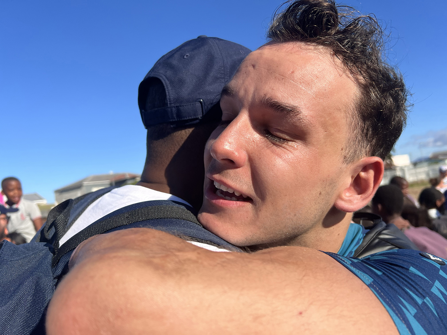 Two men embrace each other outdoors under a clear blue sky. The man facing the camera has closed eyes and a content expression, while the other man's face is not visible. People and buildings can be seen in the background.