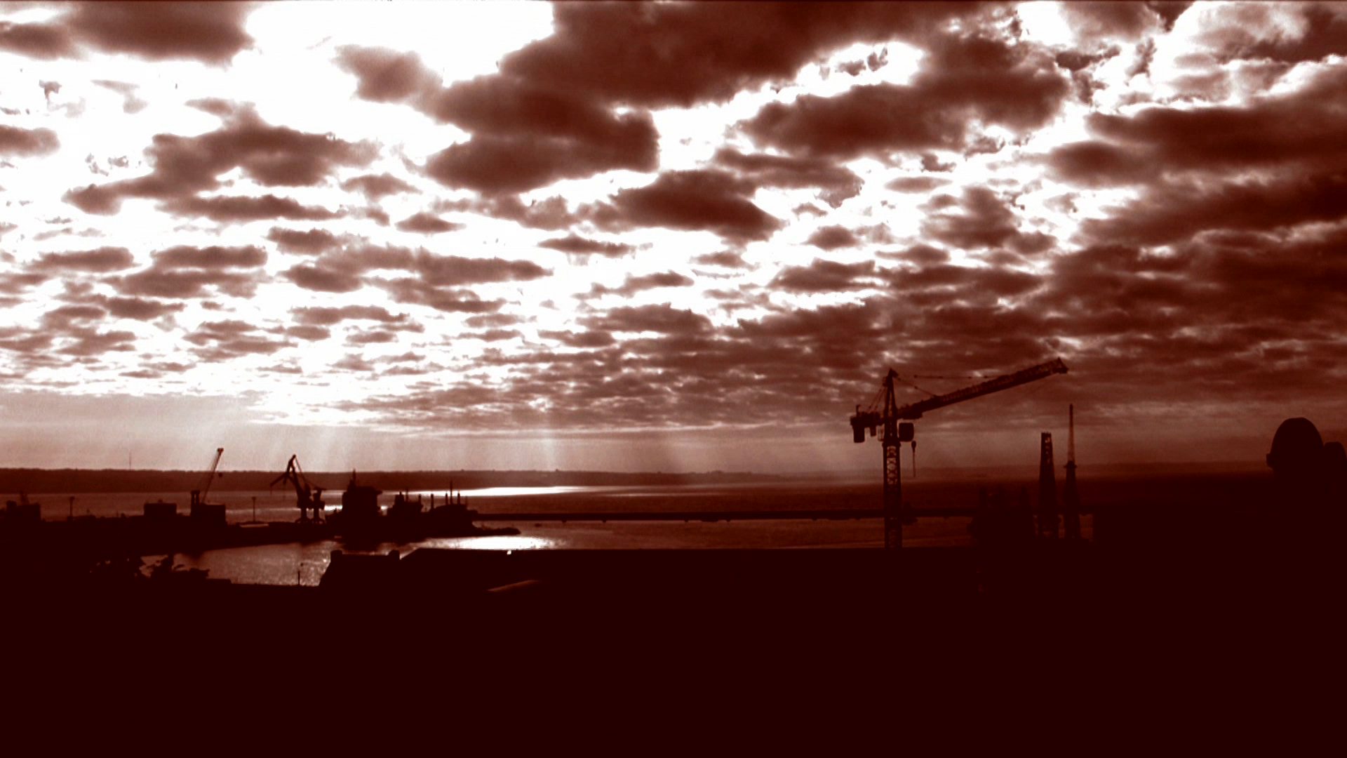 •2 Xpress2Brest_Film_Kate_Thompson_Gorry Sepia-toned image of a construction site by a waterfront. Several cranes are silhouetted against a dramatic sky filled with clouds. Sun rays peek through the clouds, casting reflections on the water below. The overall scene has an industrial yet serene atmosphere.