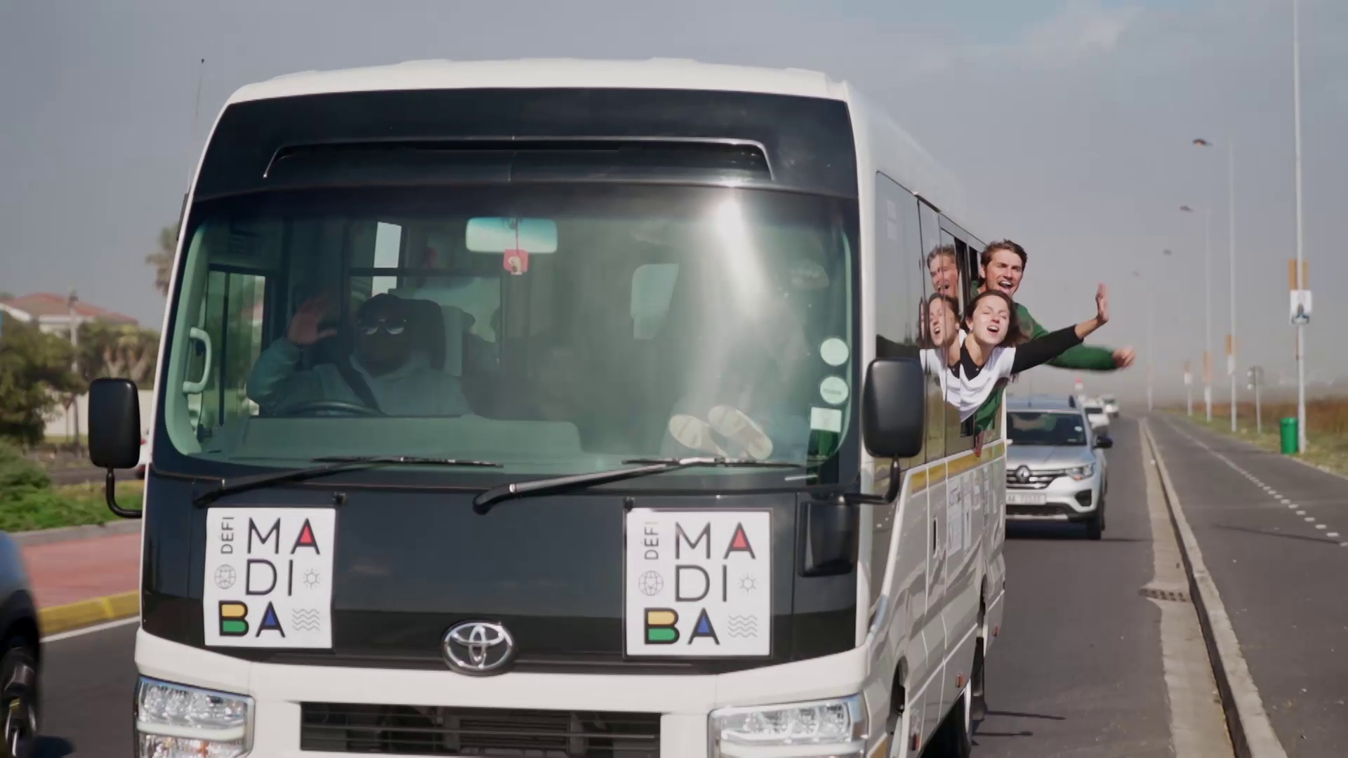 A group of joyful people lean out of the windows of a moving mini-bus, waving and smiling. The mini-bus is driving on a sunny road and has "DEFI MADIBA" banners on its front. Another vehicle is visible behind the mini-bus.