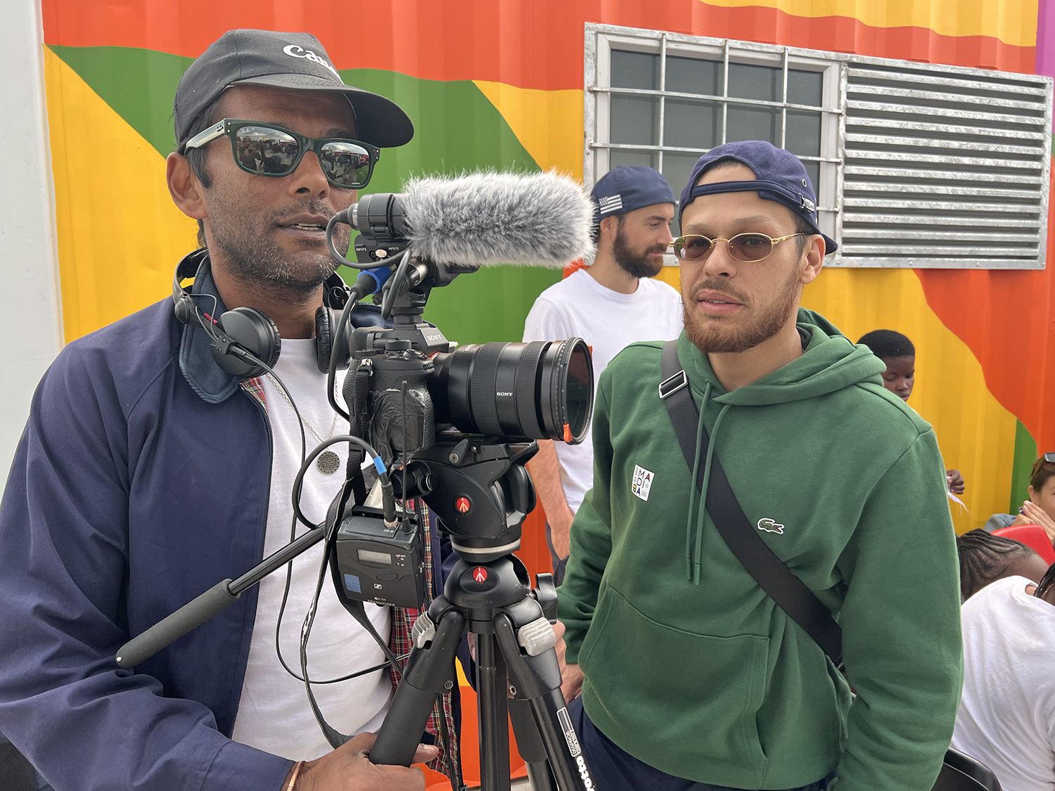 Two men, one wearing headphones and operating a camera on a tripod with a microphone, the other wearing glasses and a green hoodie, stand together. They are in front of a colorful, geometric striped background with a window. Other individuals can be seen in the background.