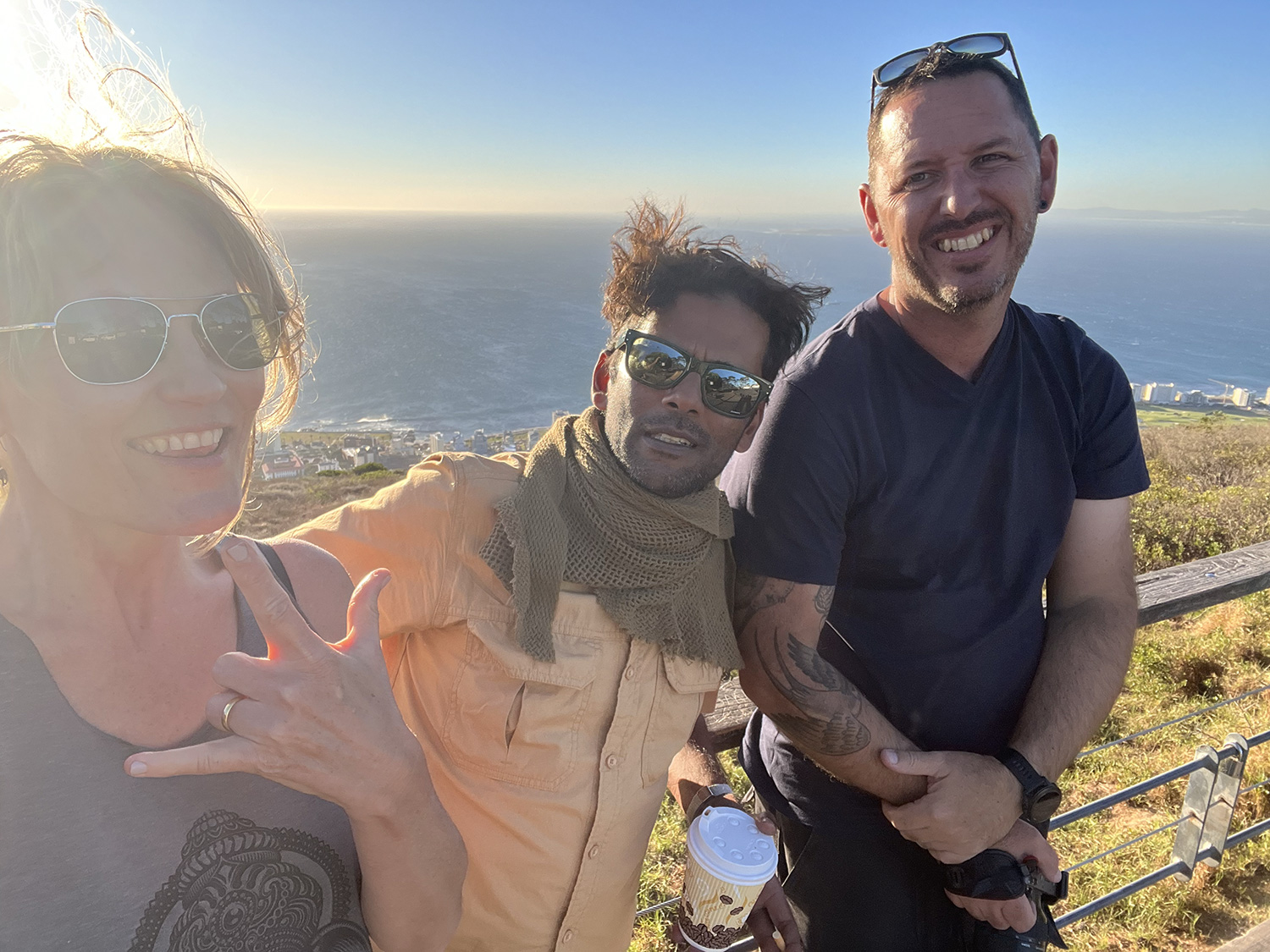 Three people, two men and one woman, pose happily on a sunny hillside overlooking a distant ocean. The woman on the left is wearing sunglasses and making a hand gesture; the man in the middle has a scarf and sunglasses; the man on the right is smiling, holding a cup.