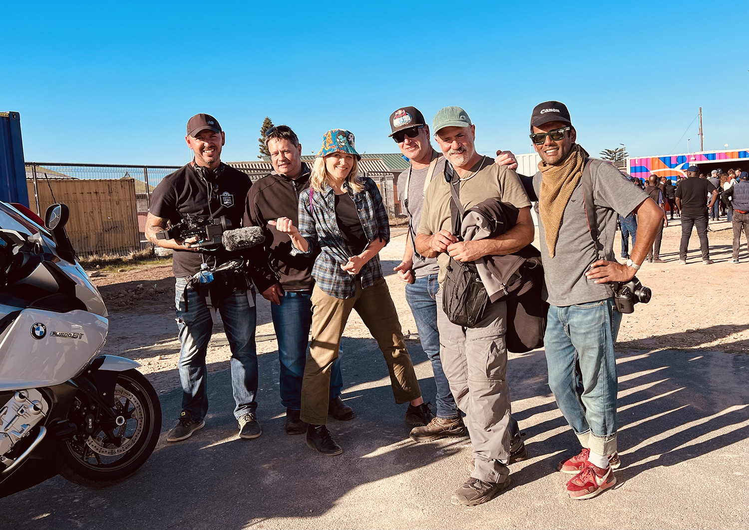 A group of six people stand outdoors smiling at the camera. They appear to be part of a film or photography crew, with some holding a camera and other equipment. A motorcycle is partially visible on the left, and the sky is clear and blue in the background.