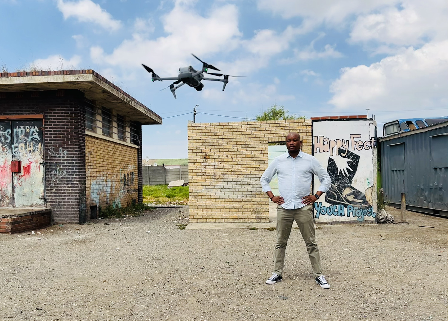 A man stands confidently with his hands on his hips in an urban area with graffiti-covered walls. A drone hovers in the air nearby. The background includes a dilapidated brick structure and a mural that reads "Happy Feet Youth Project.