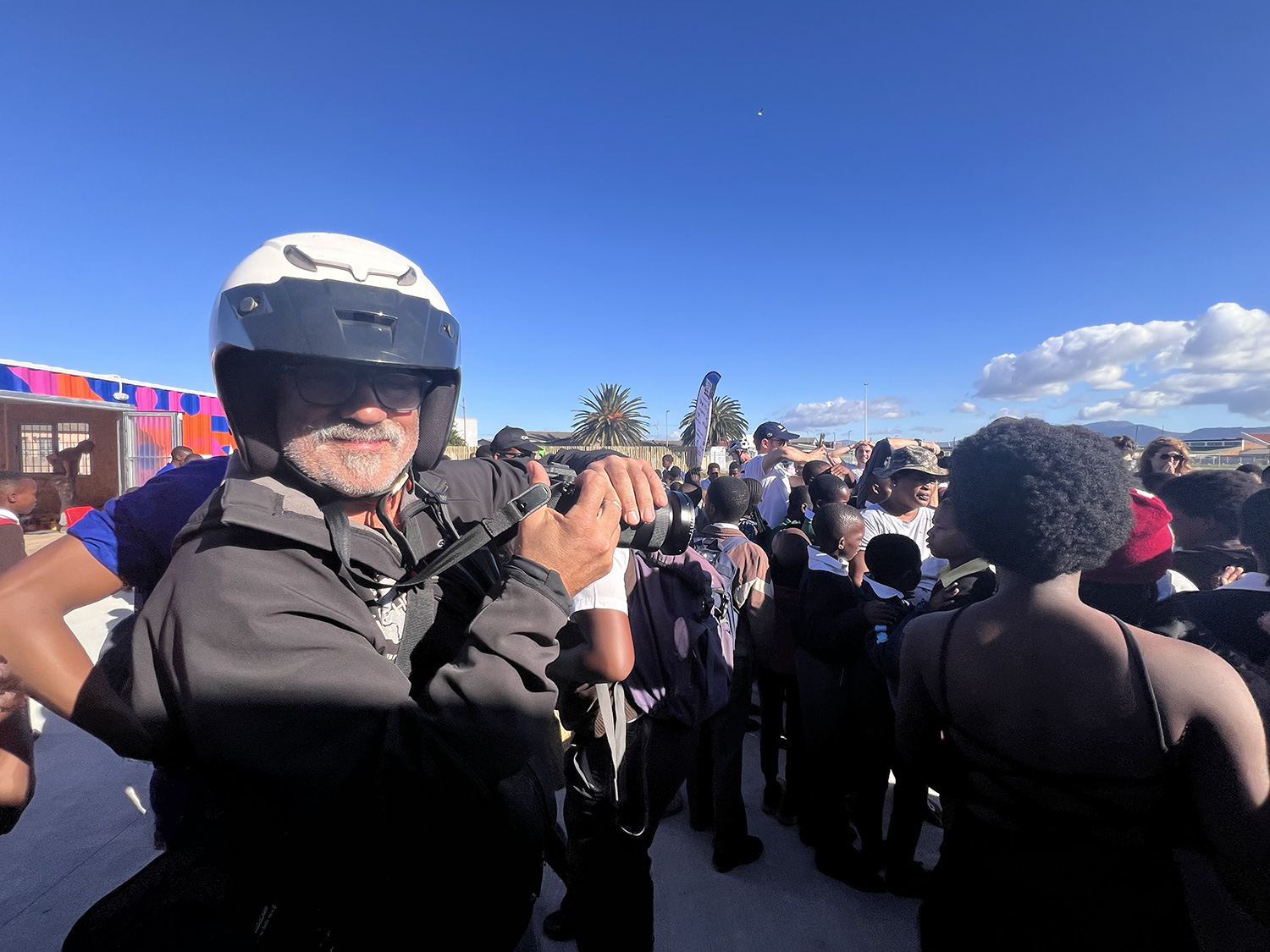 A man wearing a helmet and glasses holds a camera surrounded by a group of people. The crowd is outdoors with a palm tree and a bright blue sky with some clouds in the background.
