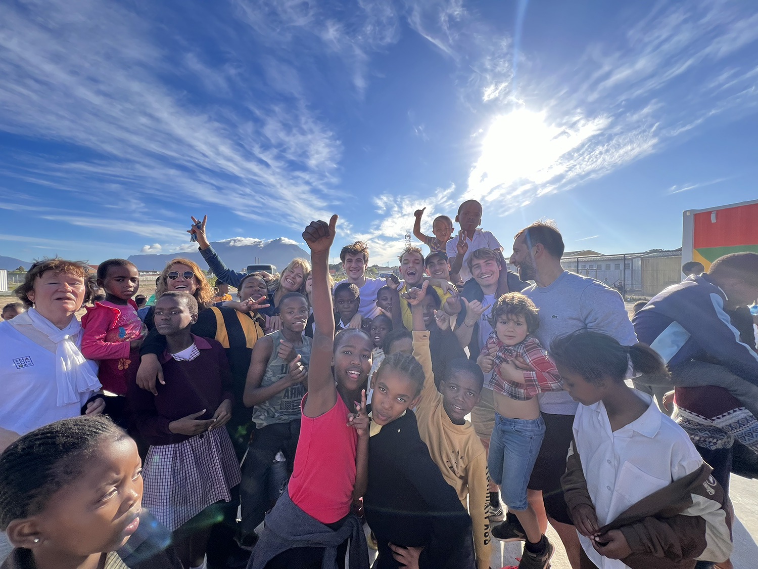 A diverse group of people, including adults and children, gather closely together outdoors under a bright, sunny sky with scattered clouds. Some individuals raise their arms in celebration, while others smile and pose for the photo.