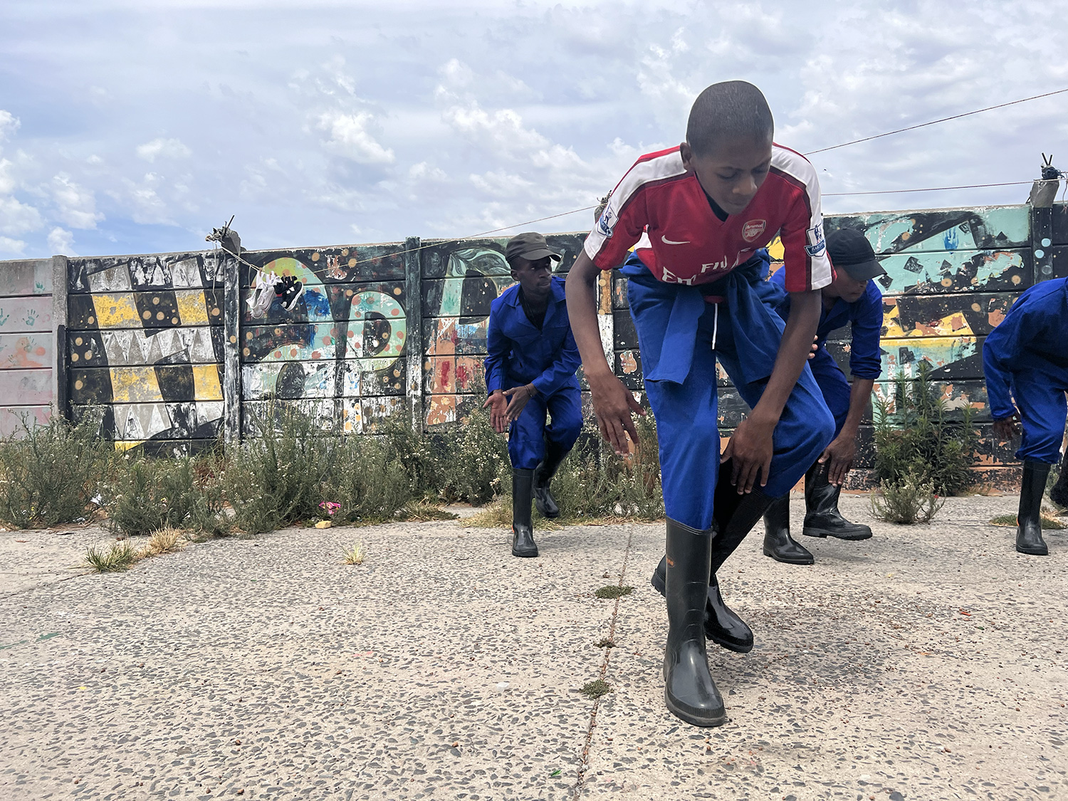 Three people dressed in blue workwear and black rubber boots are dancing outside on a concrete surface with colorful graffiti-covered walls in the background. One person, in a red shirt, is in the foreground, crouching with a focused expression.
