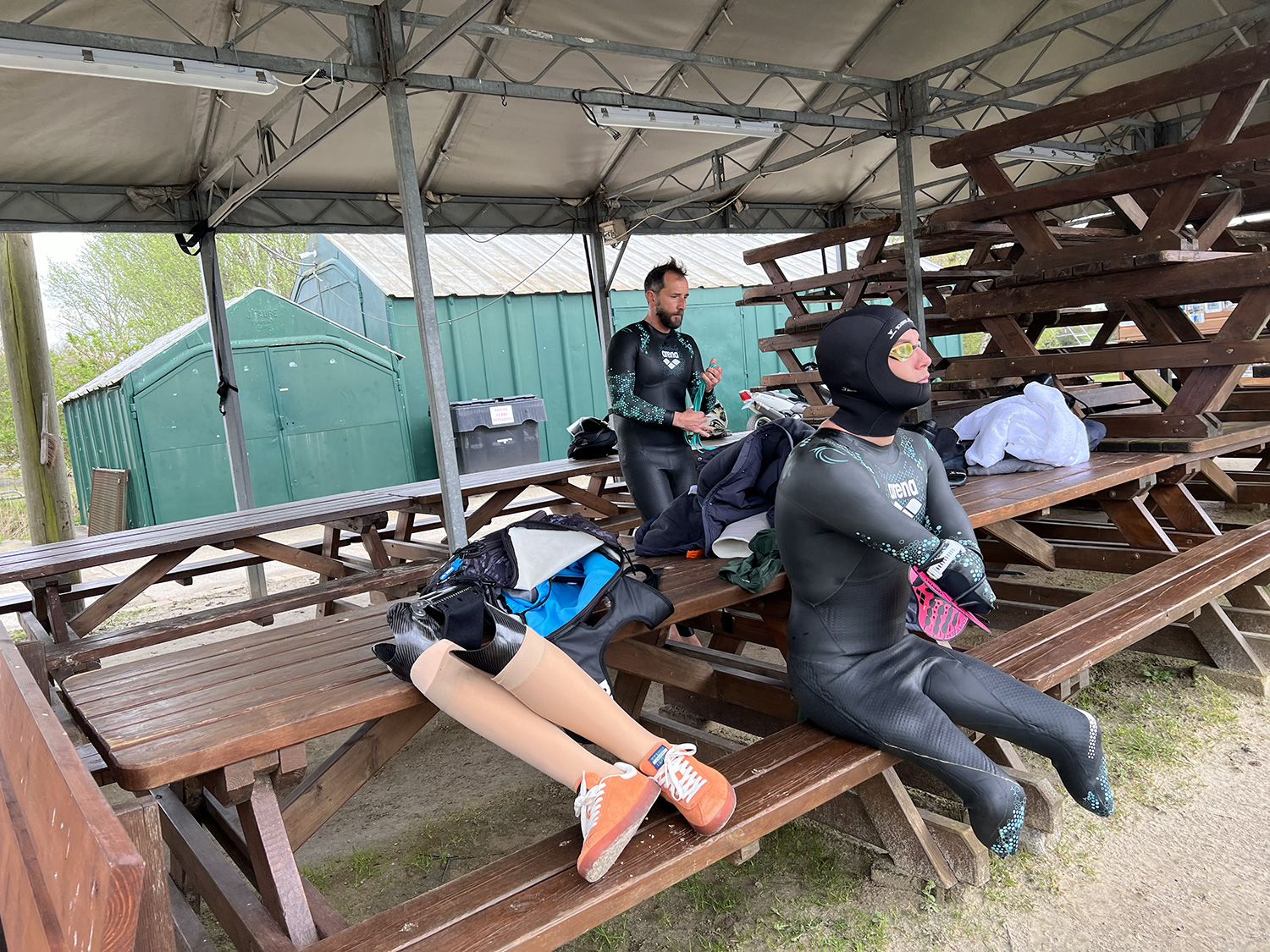 Two people in wetsuits sit on wooden picnic tables under a canopy. One person uses prosthetic legs that are removed and placed on the table nearby. The other person looks slightly away, holding sports gear. The area appears to be an outdoor recreation or training facility.