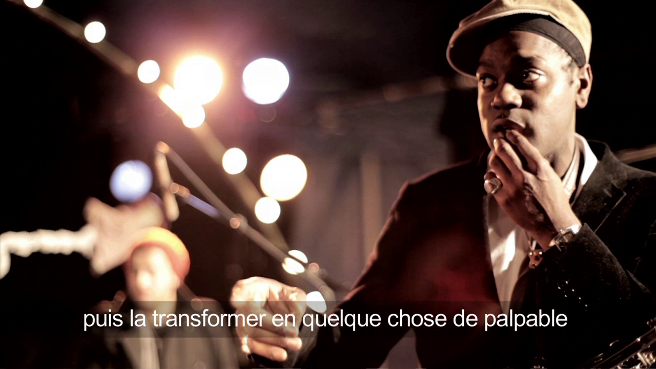 •5 Xpress2Brest_Film_Kate_Thompson_Gorry A man in a suit and hat is speaking into a microphone on stage with bright lights in the background. The French text on the image reads: "puis la transformer en quelque chose de palpable.