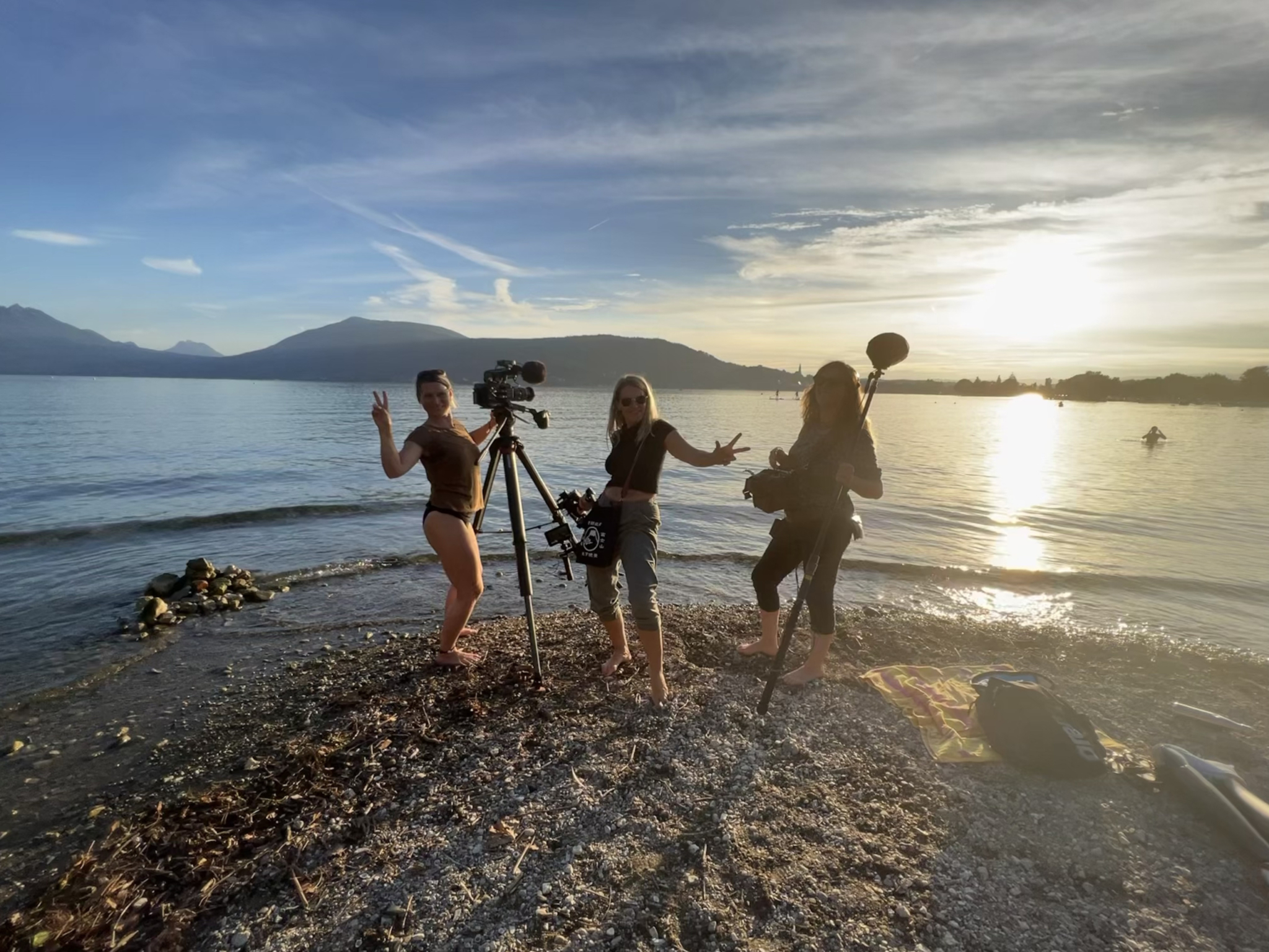 Three people stand on a rocky shore by a calm lake during sunset, one holding a boom microphone, another with a camera on a tripod, and the third with a handheld camera. They are smiling and making peace signs, enjoying the serene scenery. Mountains are in the background.
