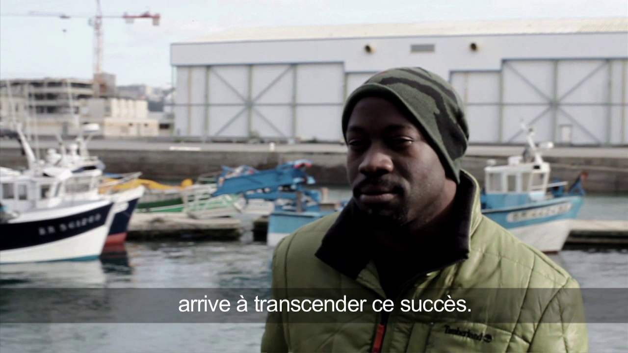 •6 Xpress2Brest_Film_Kate_Thompson_Gorry A person wearing a green jacket and beanie stands near a working harbor with boats and industrial buildings in the background. French text overlay reads: "arrive à transcender ce succès.