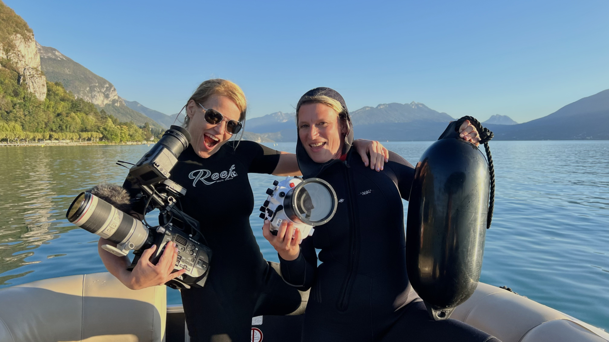 Two people on a boat wearing wetsuits and smiling, one holding a professional camera with a large lens, and the other holding underwater camera housing. They are surrounded by a picturesque lake with mountains in the background under a clear blue sky.