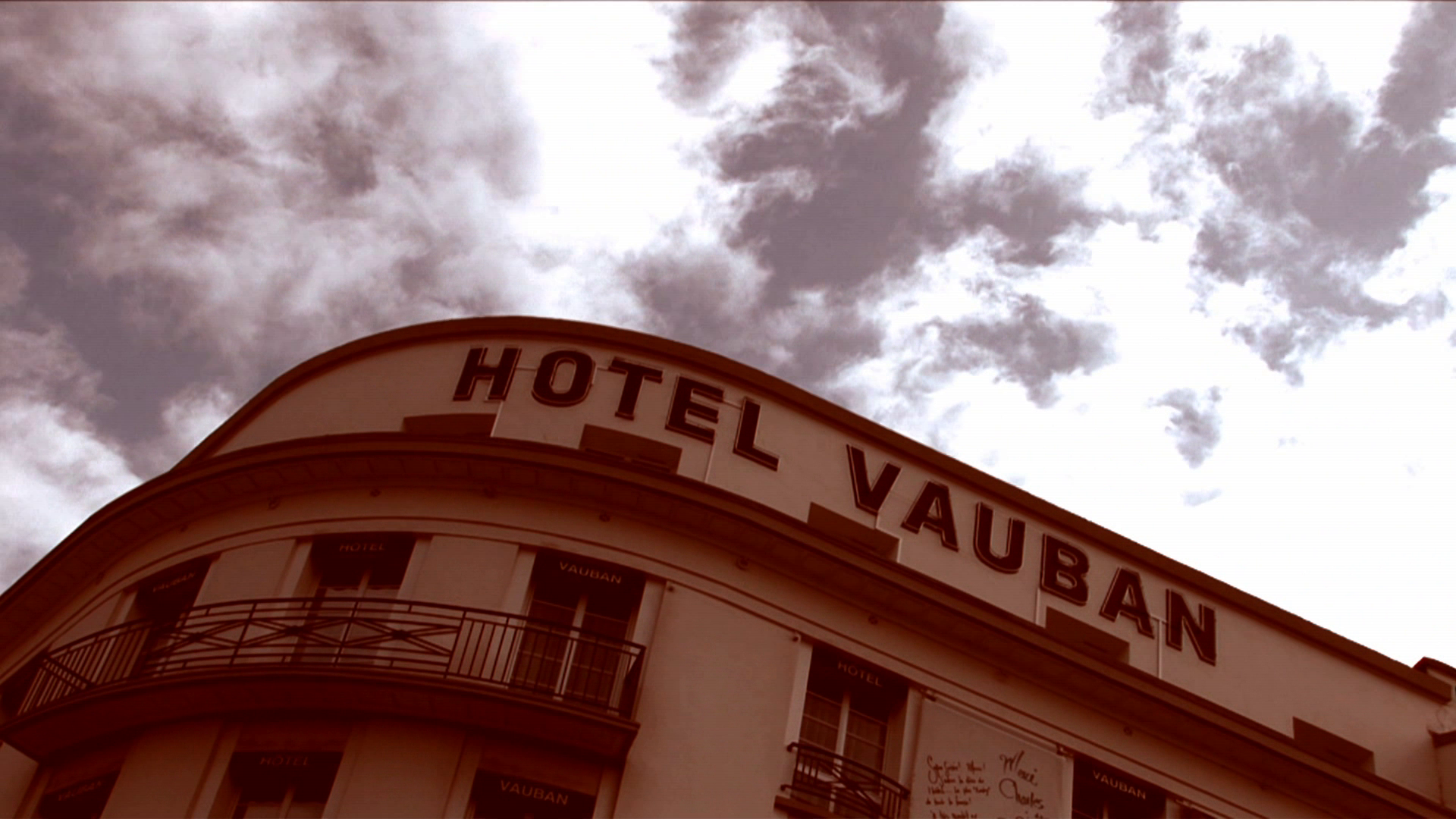 •7 Xpress2Brest_Film_Kate_Thompson_Gorry A sepia-toned photo of the upper part of a building with a sign reading "HOTEL VAUBAN" on its front facade. The sky above is partly cloudy, adding a dramatic effect to the image. The building features balconies with metal railings.