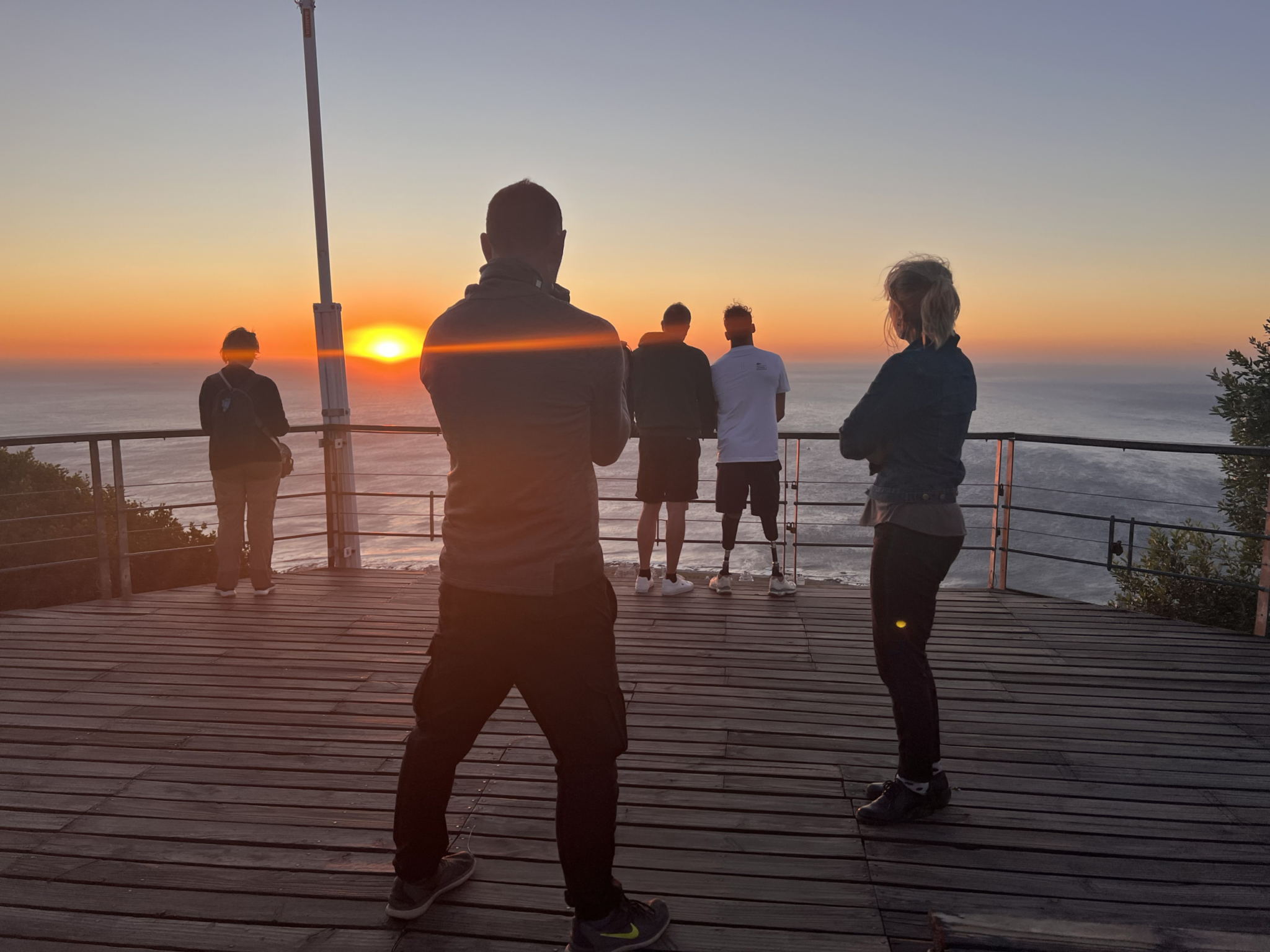 People are standing on a wooden deck at sunset, overlooking a body of water. The sun is low on the horizon, casting an orange glow. Some individuals are gazing at the sunset while others are engaging in conversation or taking photos. A railing borders the deck.