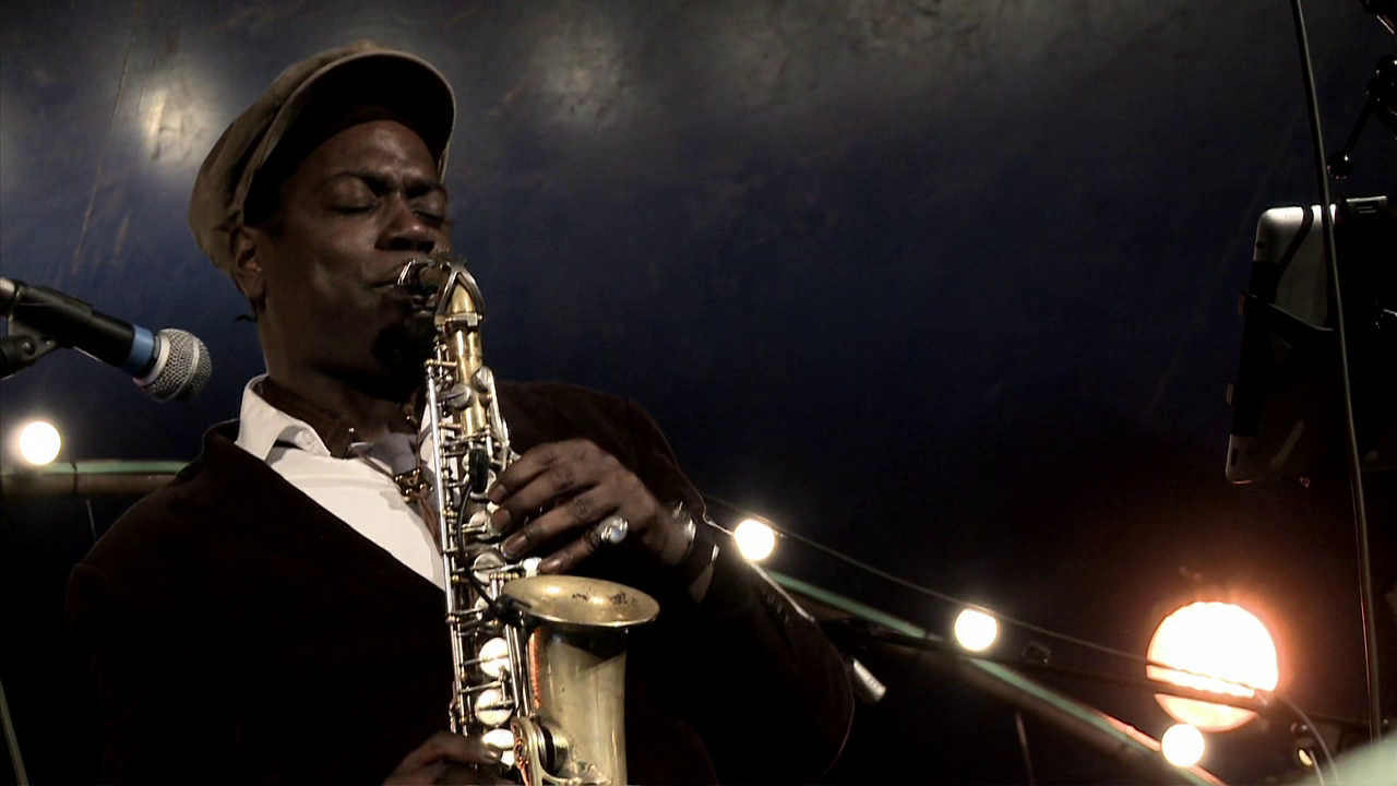 •8 Xpress2Brest_Film_Kate_Thompson_Gorry A man wearing a hat plays a saxophone passionately on a dimly lit stage. The background is dark with a few blurred, circular lights. He is focused and his eyes are closed, immersed in the music.
