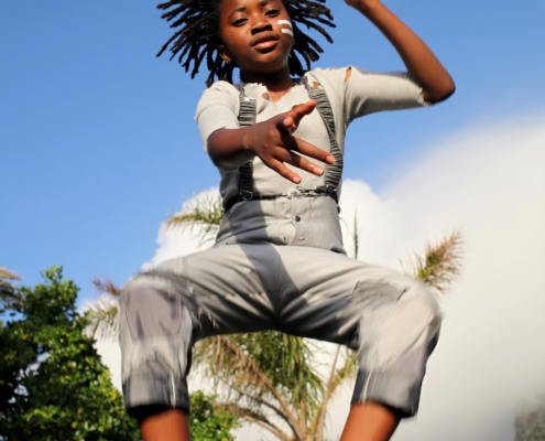 A child with dreadlocks and wearing grey overalls is captured mid-jump against a blue sky in this poster for the film “Dare to Dream, Zip Zap Circus." The text at the top displays the film’s title, while production details are listed at the bottom.
