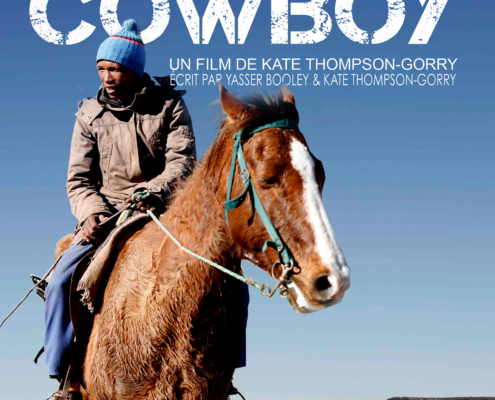 A man in rugged clothing rides a brown and white horse through a mountainous desert landscape under a clear blue sky. "Karoo Cowboy" and credits appear at the top of the image, perfectly encapsulating the spirit of Western adventure.
