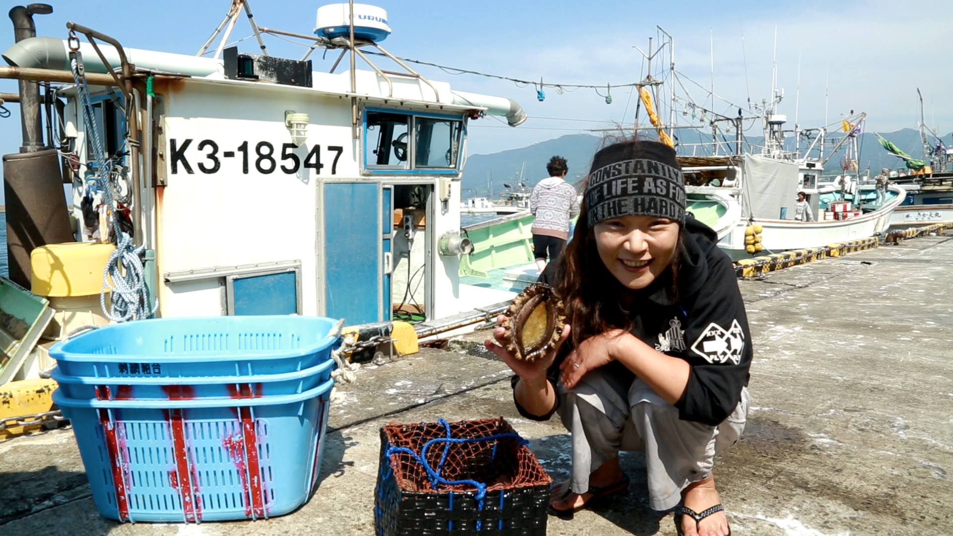 A person crouches on a pier next to a fishing boat, holding a large mollusk and smiling at the camera. They wear a beanie with text and have baskets full of fishing equipment nearby. The background reveals other boats and a mountainous landscape.
