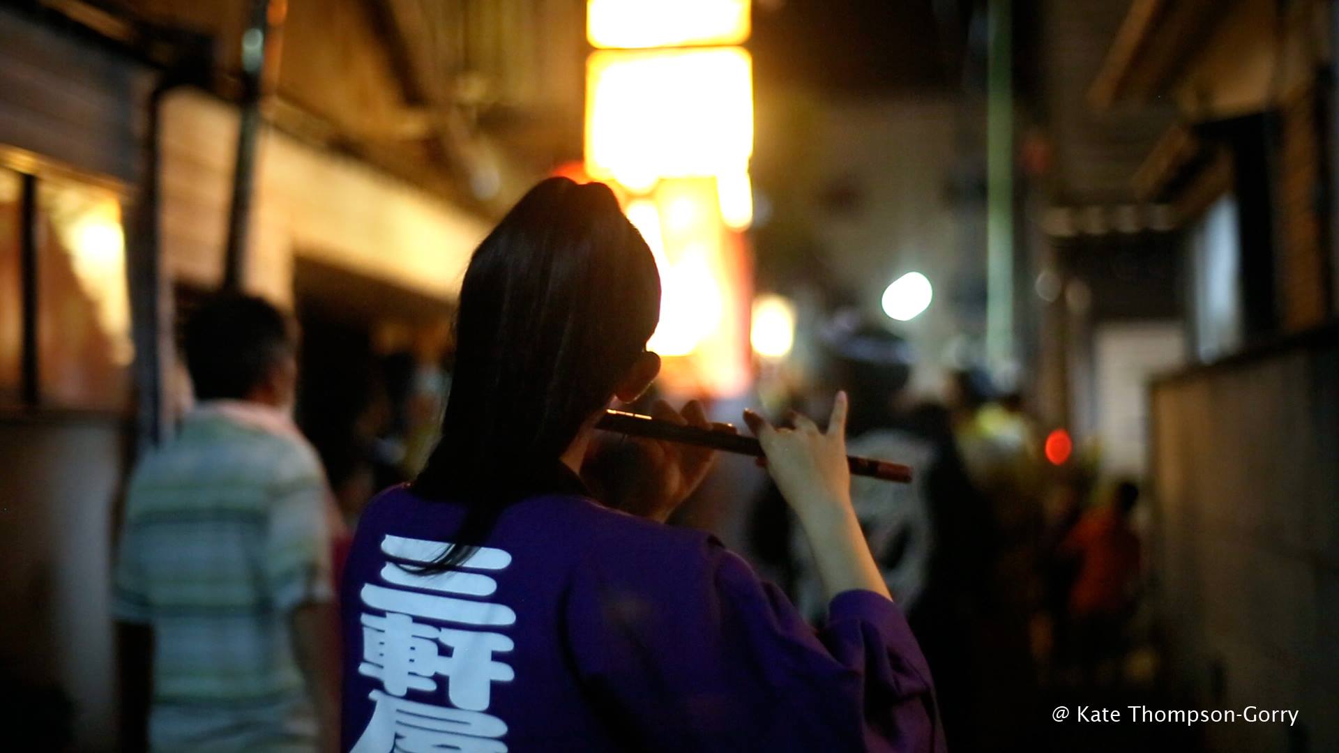 A person in a dark purple outfit plays a flute in the middle of a dimly-lit street at night. Bright lights illuminate the background and other people are barely visible around the performer. © Kate Thompson-Gorry.