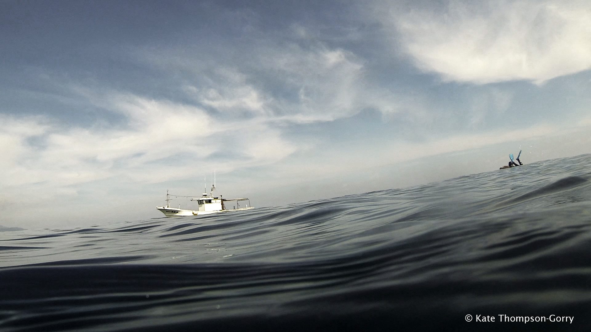 A boat floats on calm ocean waters under a partly cloudy sky. In the distance, a person can be seen swimming or snorkeling, their arms extended upward. The image captures the serene seascape and the vastness of the open water.